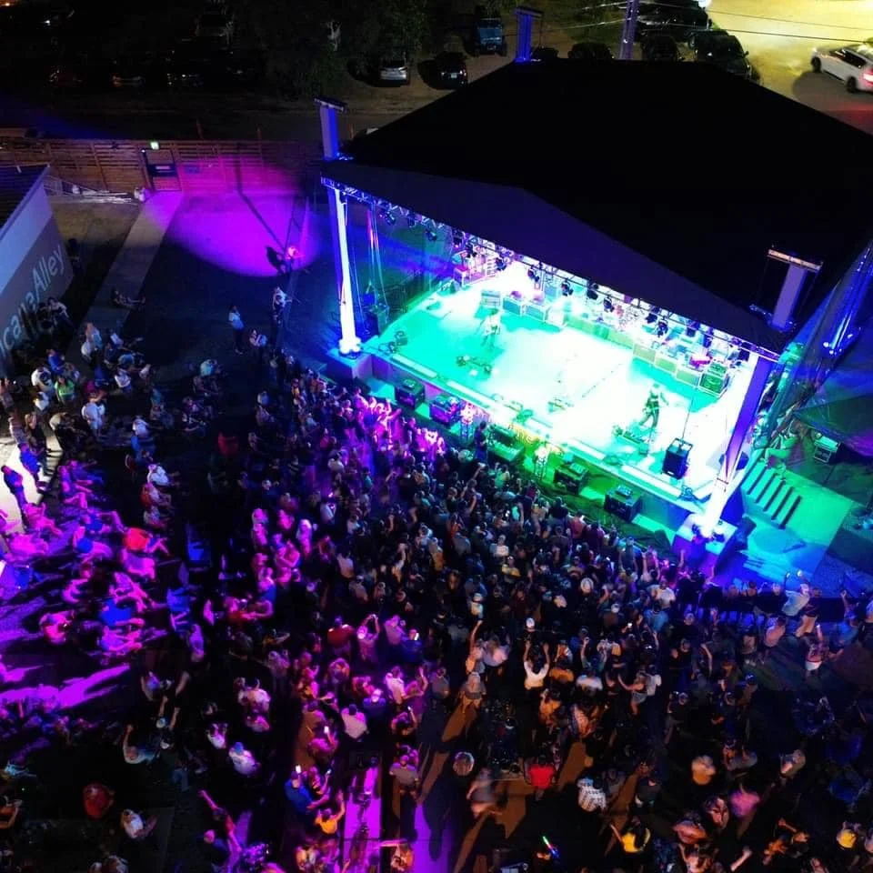An outdoor concert or event taking place on a stage with colorful lighting, with a large audience seated and standing in front of the stage, under a pavilion at night.