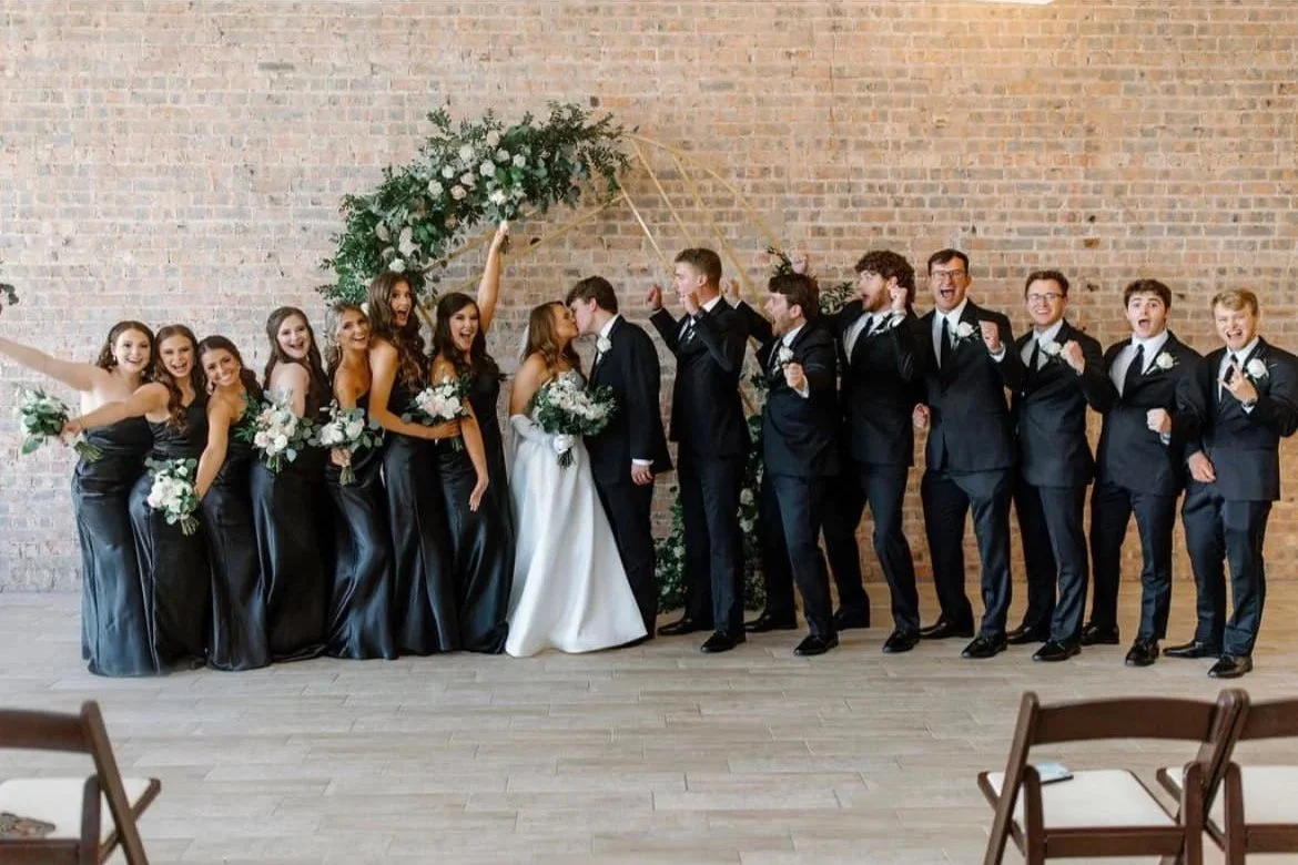 Wedding party with bride and groom kissing, surrounded by bridesmaids and groomsmen, standing under a floral arch against a brick wall.