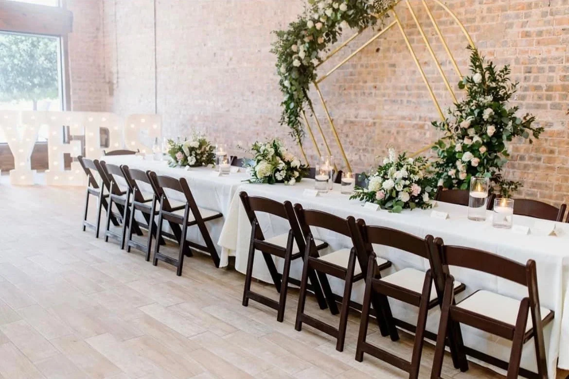 A decorated reception table with flowers, candles, and chairs, with large illuminated letters spelling 'YHPE' and a geometric floral wall art attached to a brick wall in the background.