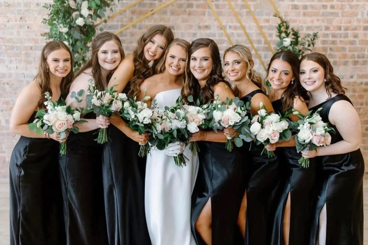 A bride and her seven bridesmaids in a wedding photo, all holding bouquets of white and pink flowers, standing in front of a brick wall with gold geometric decorations.