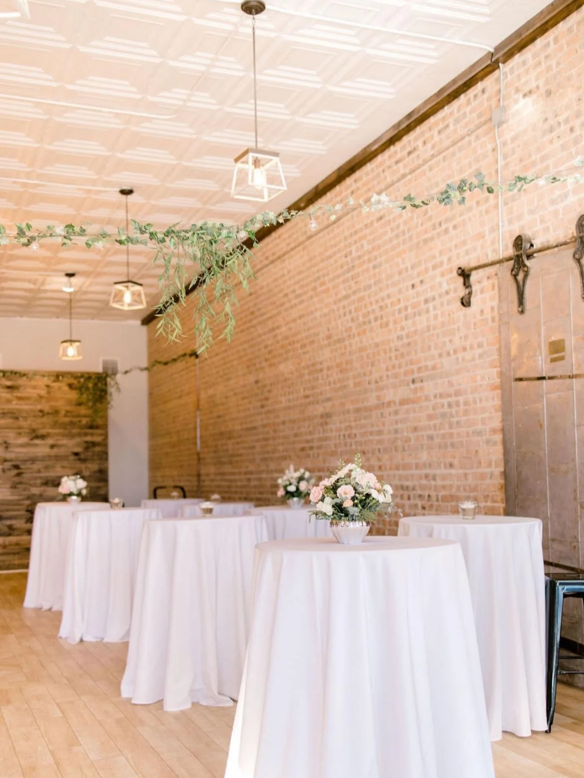 Indoor event space with wooden floors, exposed brick walls, hanging light fixtures, and decorated tables with white tablecloths and floral centerpieces.