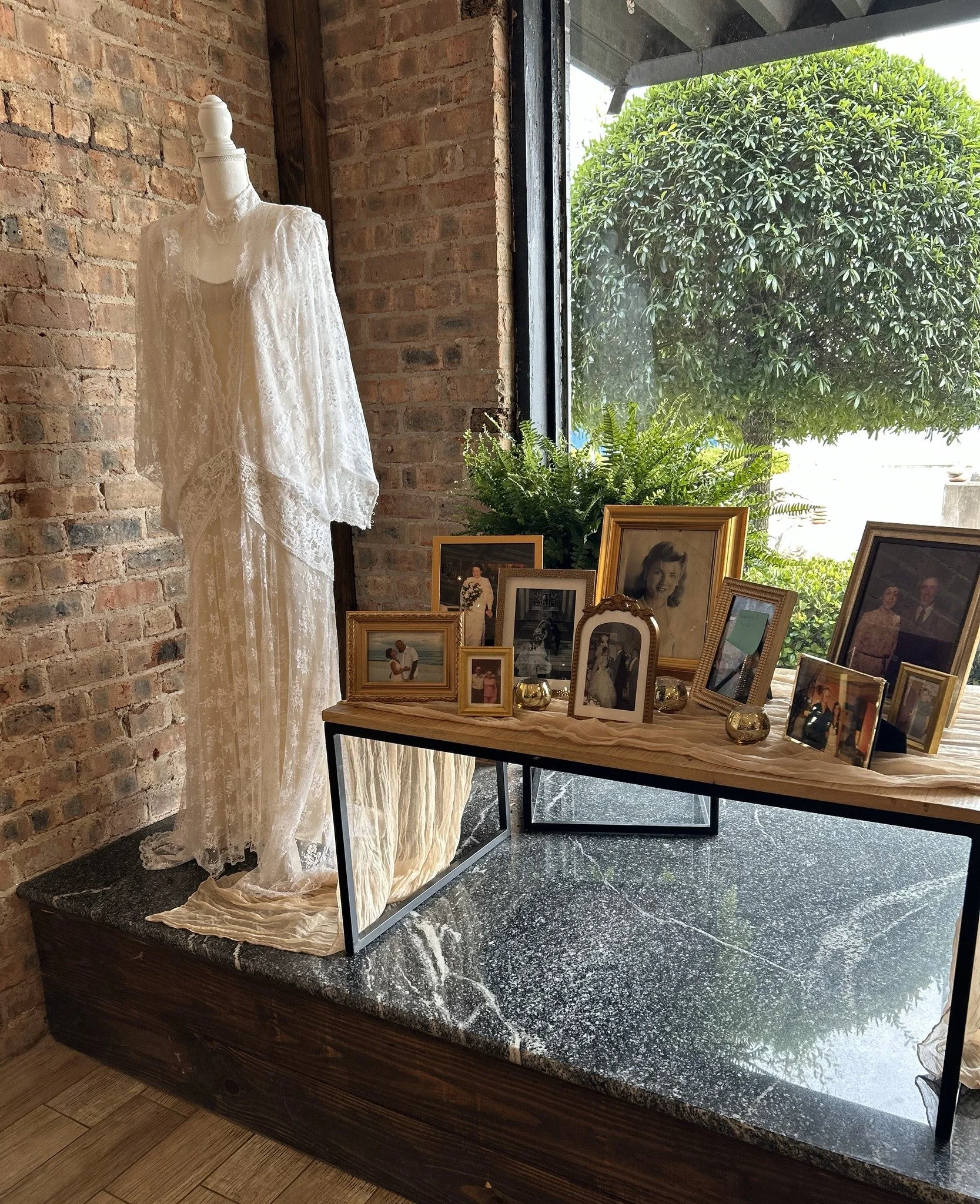 Display of vintage photographs and a white lace dress in a rustic indoor setting with exposed brick wall, wooden frame, greenery outside window, and black marble platform.