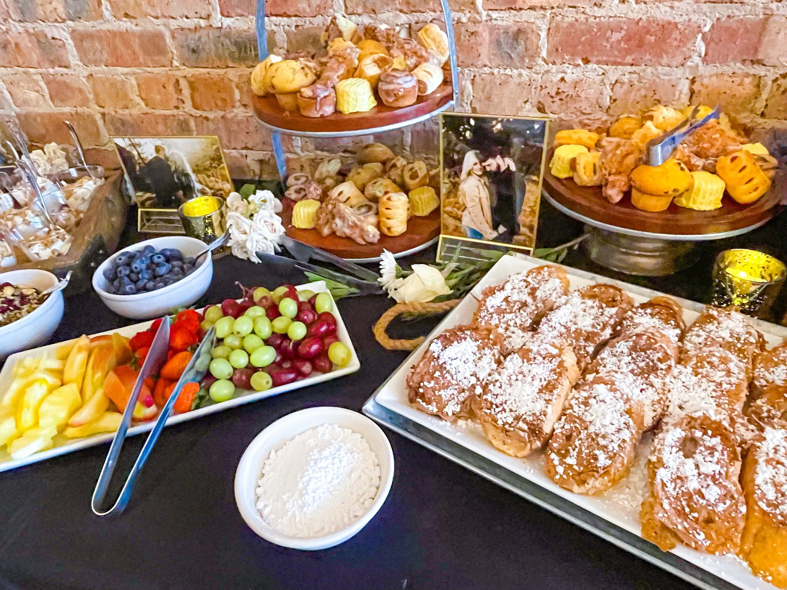 A dessert table with various pastries and fresh fruit, including a tray of powdered sugar-topped pastries, bowls of blueberries, grapes, and melon, and a setup with photos and decorative items against a brick wall.