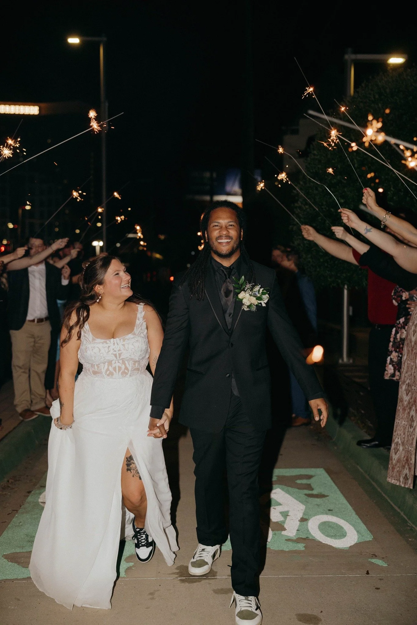 A happy couple, a bride in a white dress and a groom in a black suit, walking hand in hand during a nighttime celebration surrounded by friends holding sparklers.