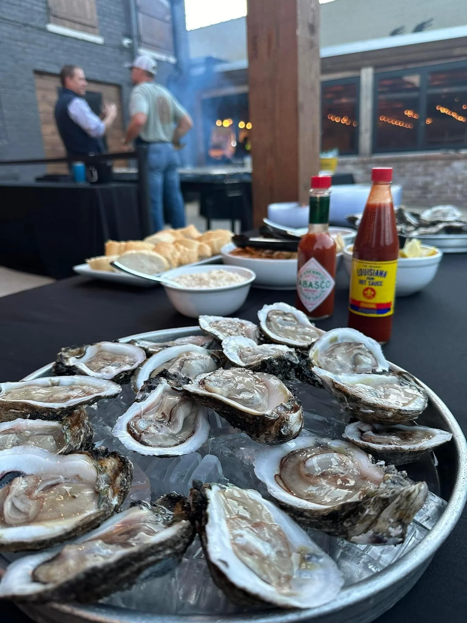 Fresh oysters on a bed of ice at a seafood dinner setup with hot sauce, cocktail sauce, rice, and bread in the background, with a grill and people in conversation in an outdoor dining area.