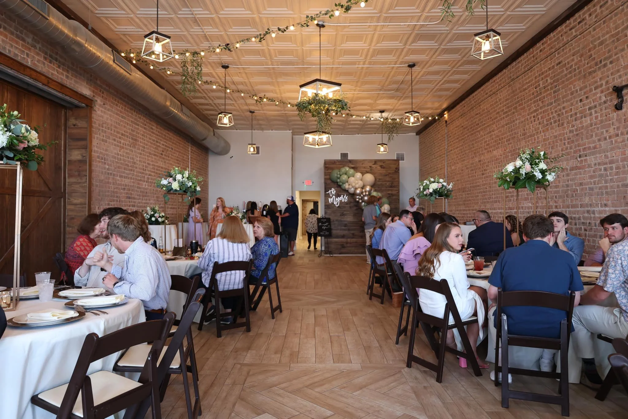 People seated at tables with white tablecloths in a decorated event space with brick walls, wooden floors, and hanging light fixtures, attending a social gathering or celebration.