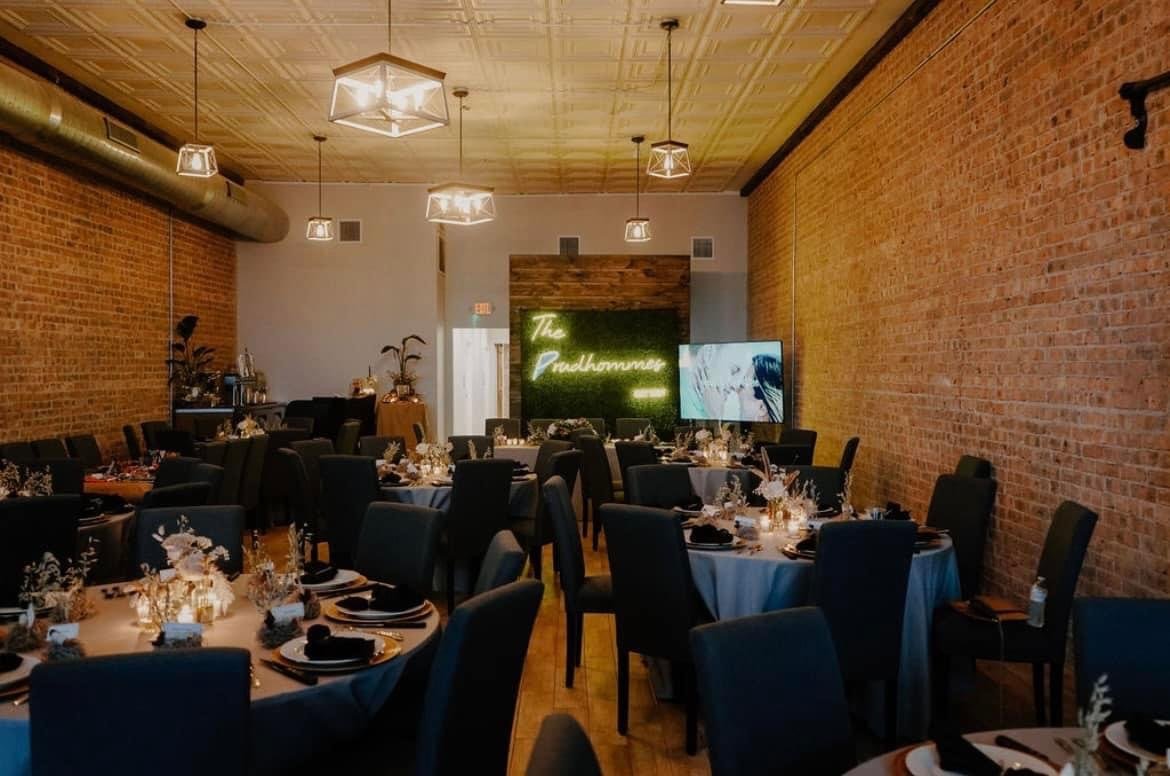Restaurant or event space with round tables set with black napkins and decorative centerpieces, black chairs, exposed brick walls, wood ceiling, and a neon sign reading 'The Pulhammery' on a green wall with a flat-screen TV nearby.