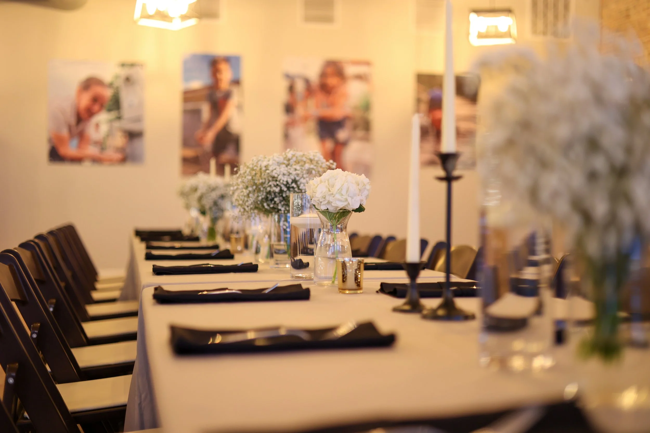 A long dining table decorated with white flowers and black napkins, set up for an event in a warmly lit room with artwork on the walls.
