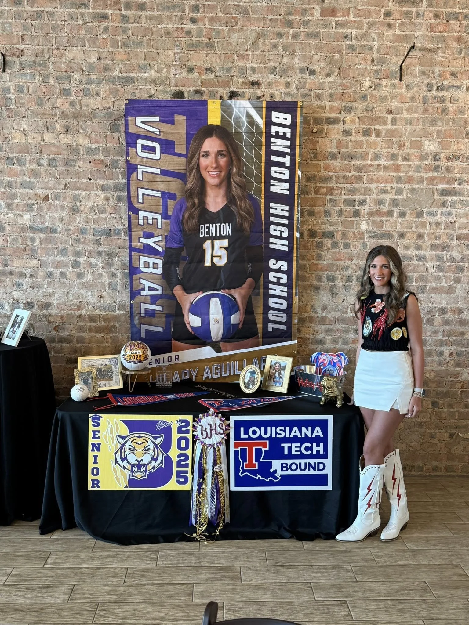 A young woman stands next to a table displaying a college commitment for Louisiana Tech and a senior banner for Lula Lady Aguilar. The table has various framed photographs, a basketball, and decorative items. A large poster behind her features a fema