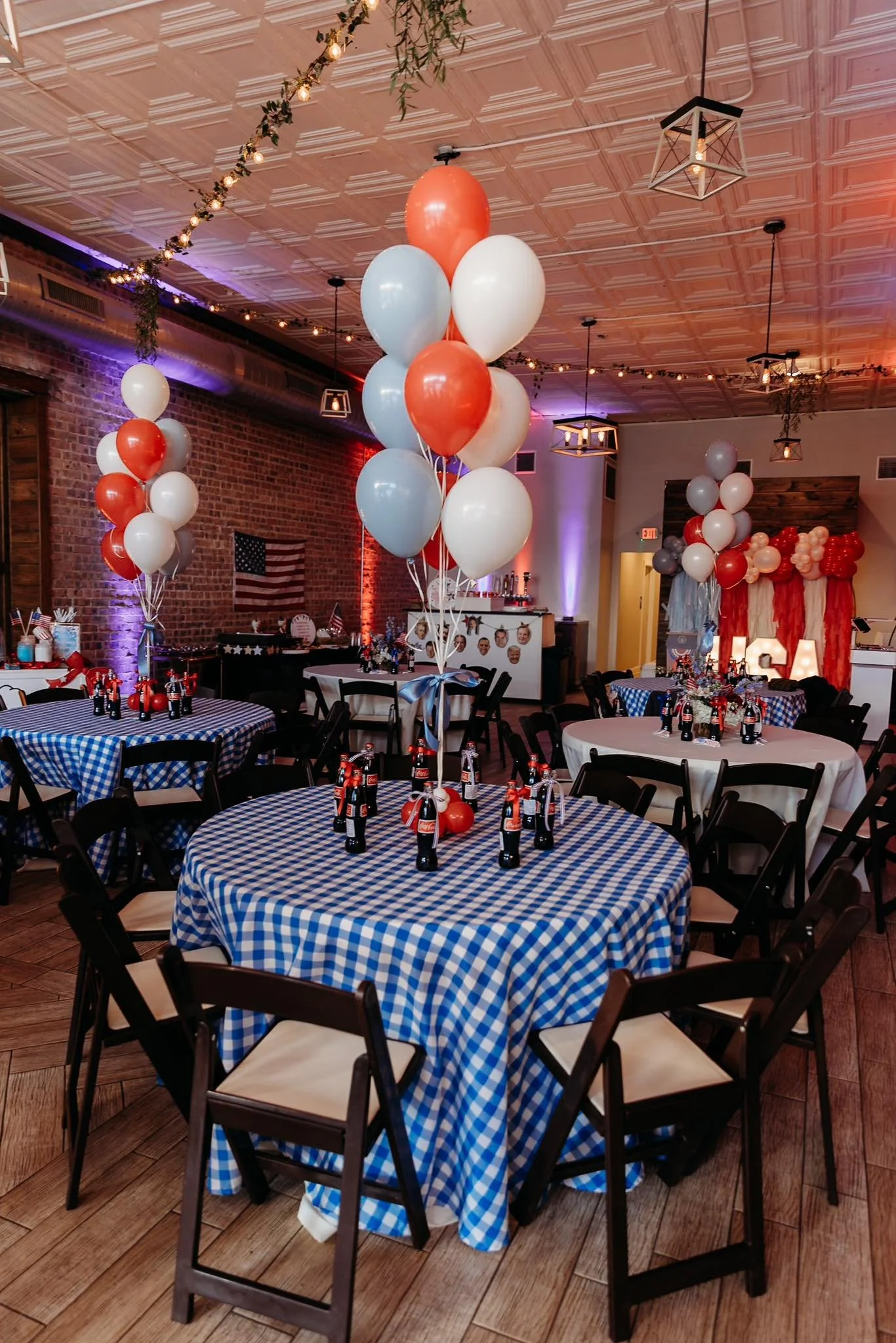 Decorated indoor event space with round tables covered in blue and white checkered tablecloths, black chairs, and balloons in red, white, and blue. The ceiling has string lights, and the background features a brick wall with an American flag and a wa