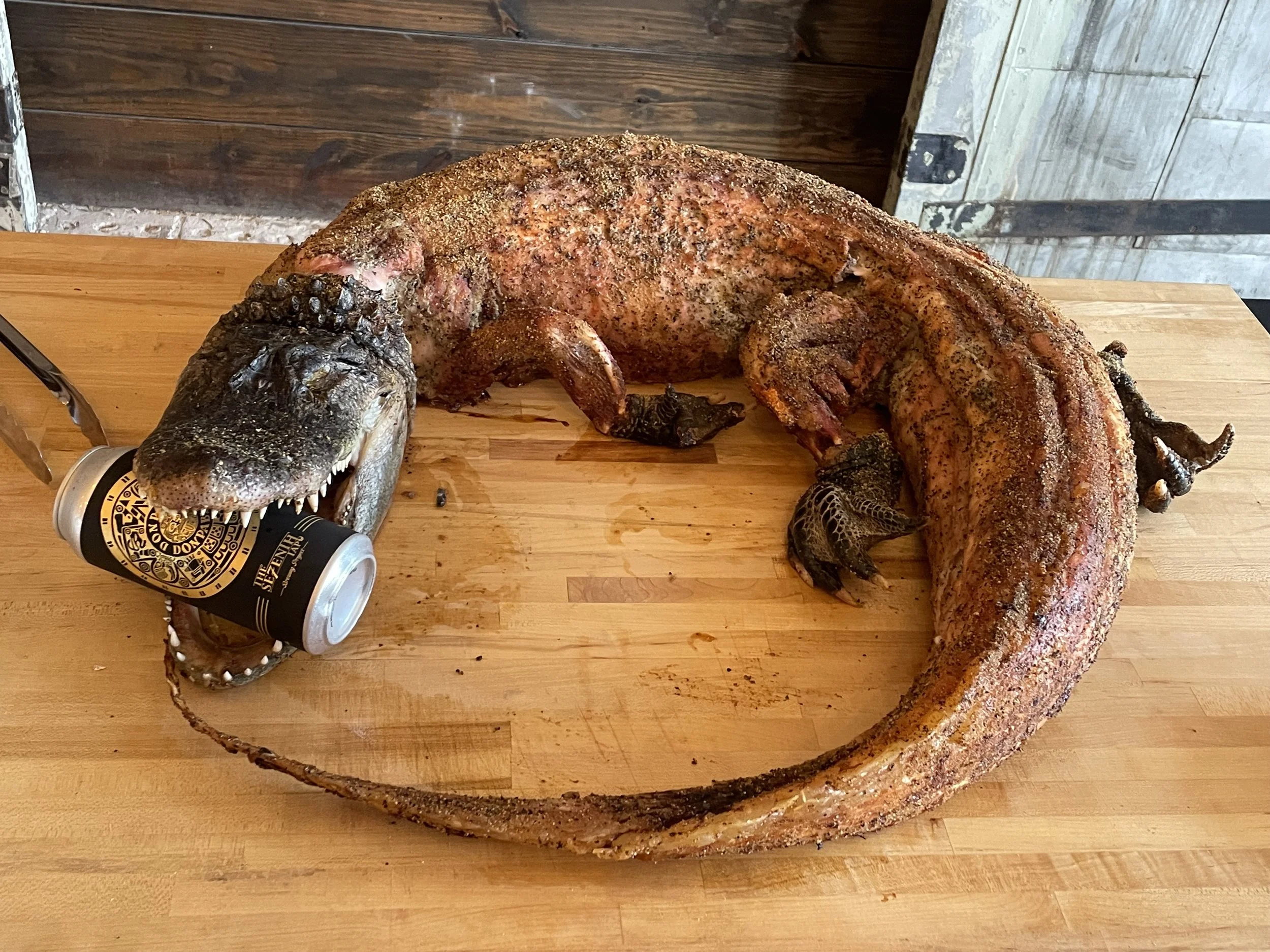A crocodile-shaped cake or sculpture holding a beer can in its mouth, placed on a wooden surface.