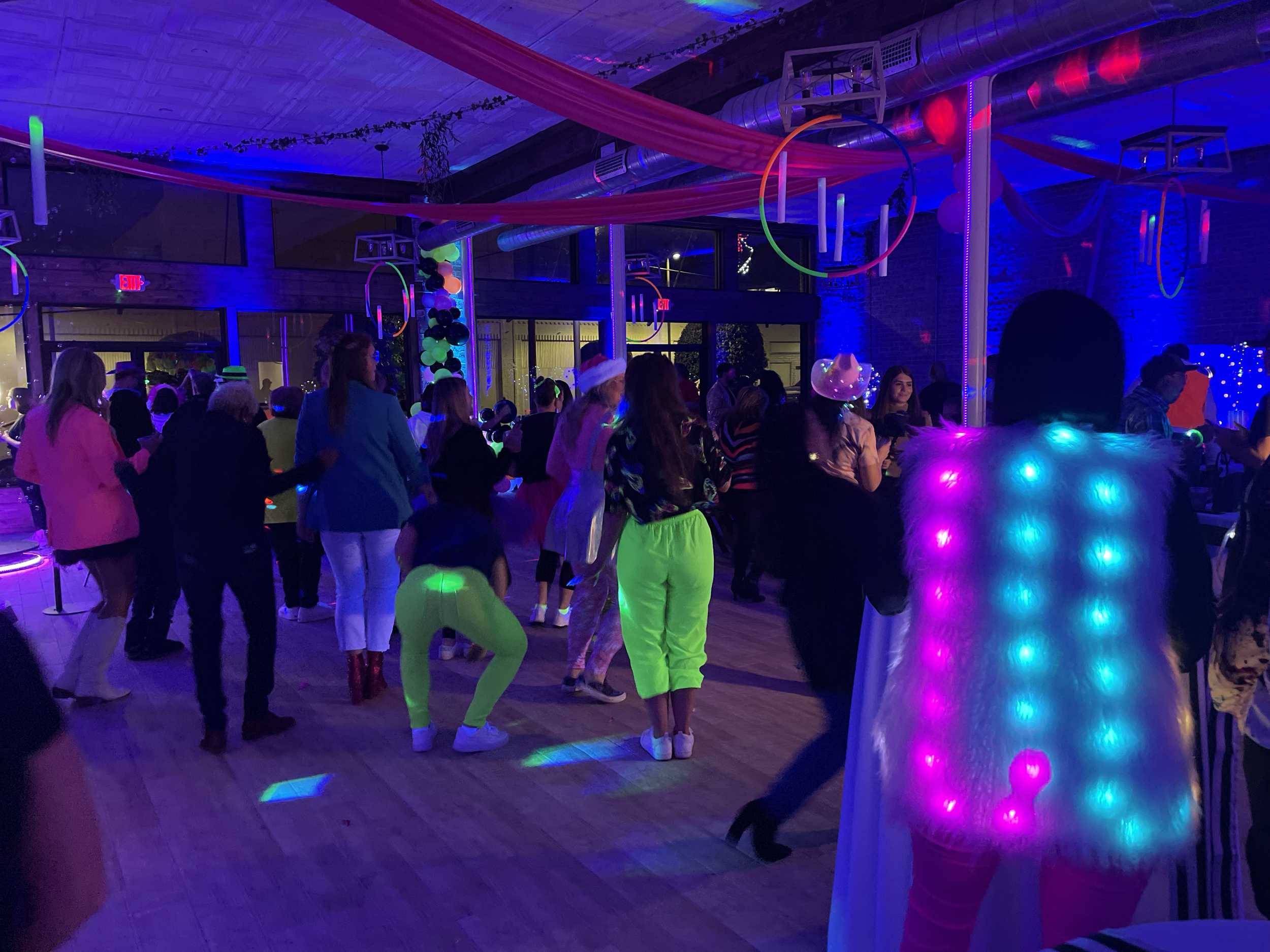 People dancing at a party with colorful glow-in-the-dark lights, decorations hanging from the ceiling, and some attendees wearing costumes and glow accessories.