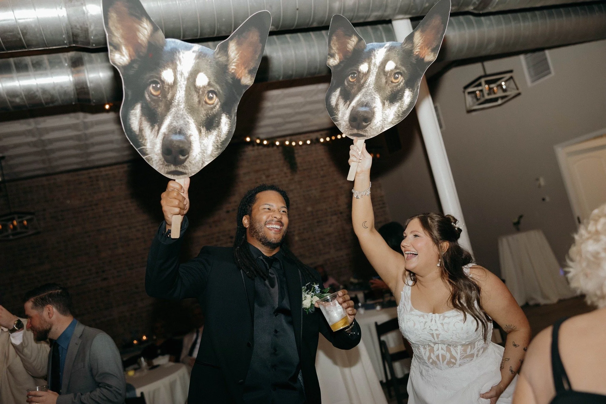 A wedding reception with a man and woman holding dog face masks on sticks, smiling and celebrating.
