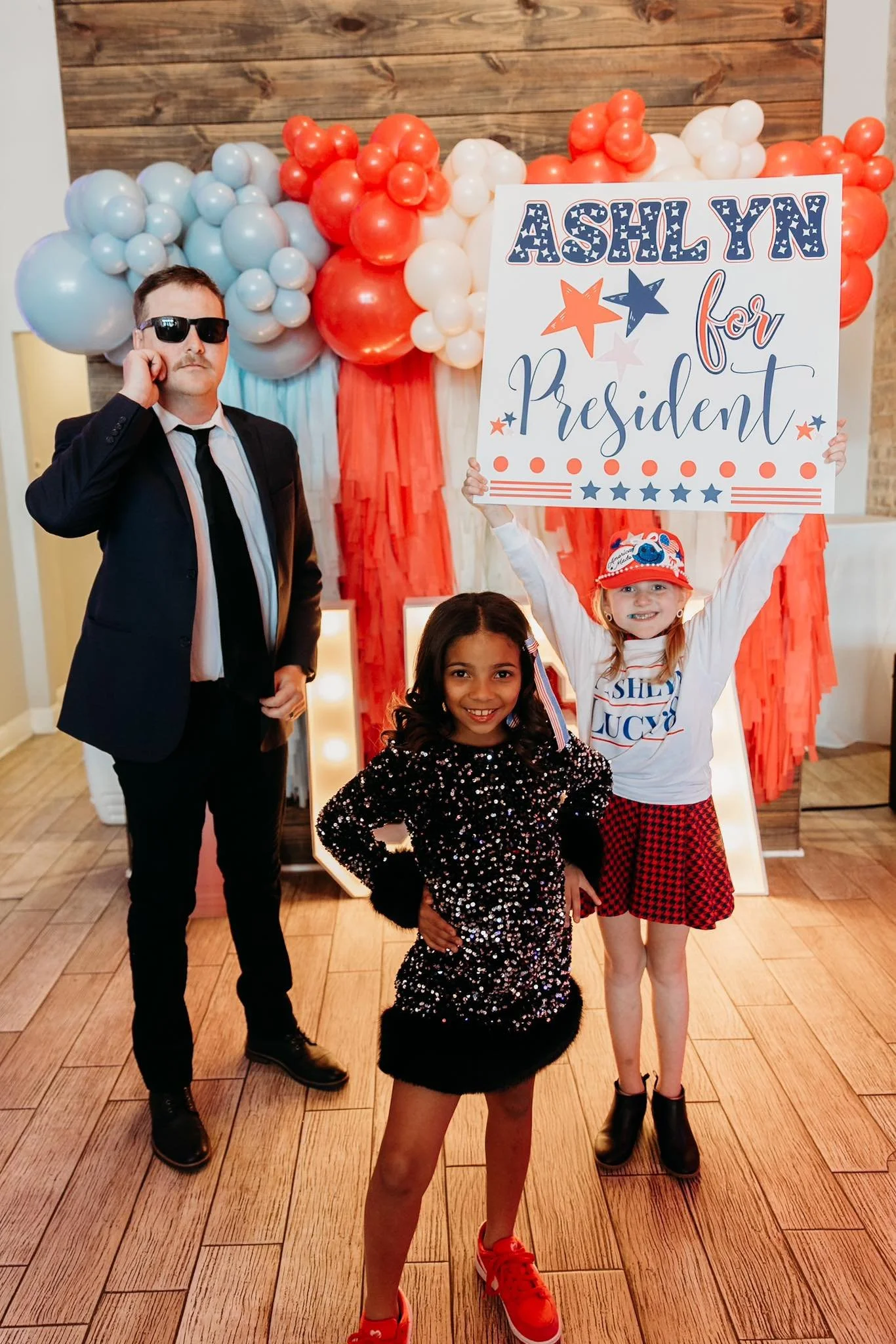Three children posing in front of a patriotic backdrop with balloons and a sign. The girl in the front wears a black sequined dress and red sneakers, smiling with her hands on her hips. The girl in the back holding the sign wears a white shirt, red a