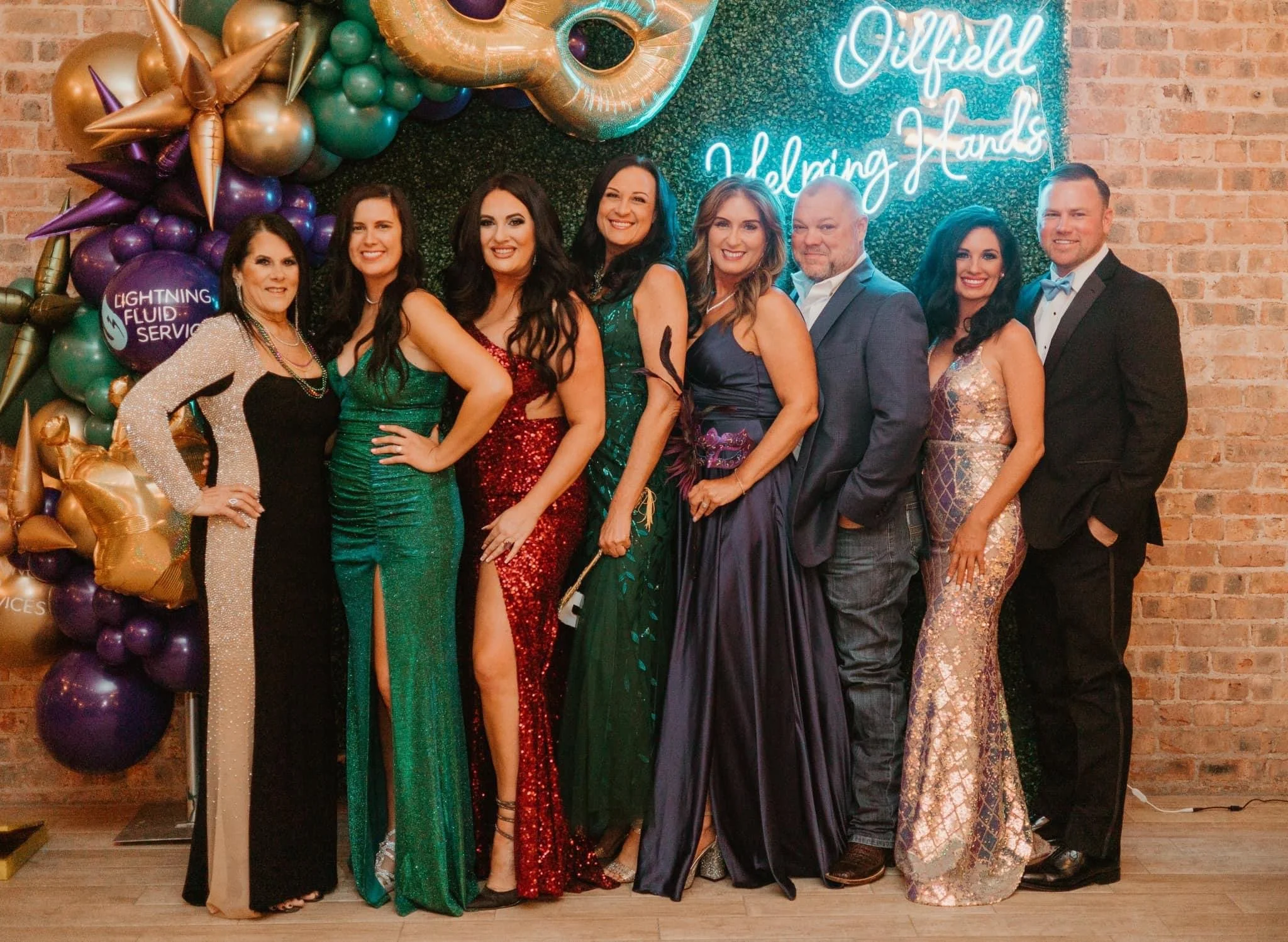 A group of ten people dressed in formal attire posing in front of a decorated party backdrop with balloons and a neon sign that reads 'Dixfield Holding Hands'.