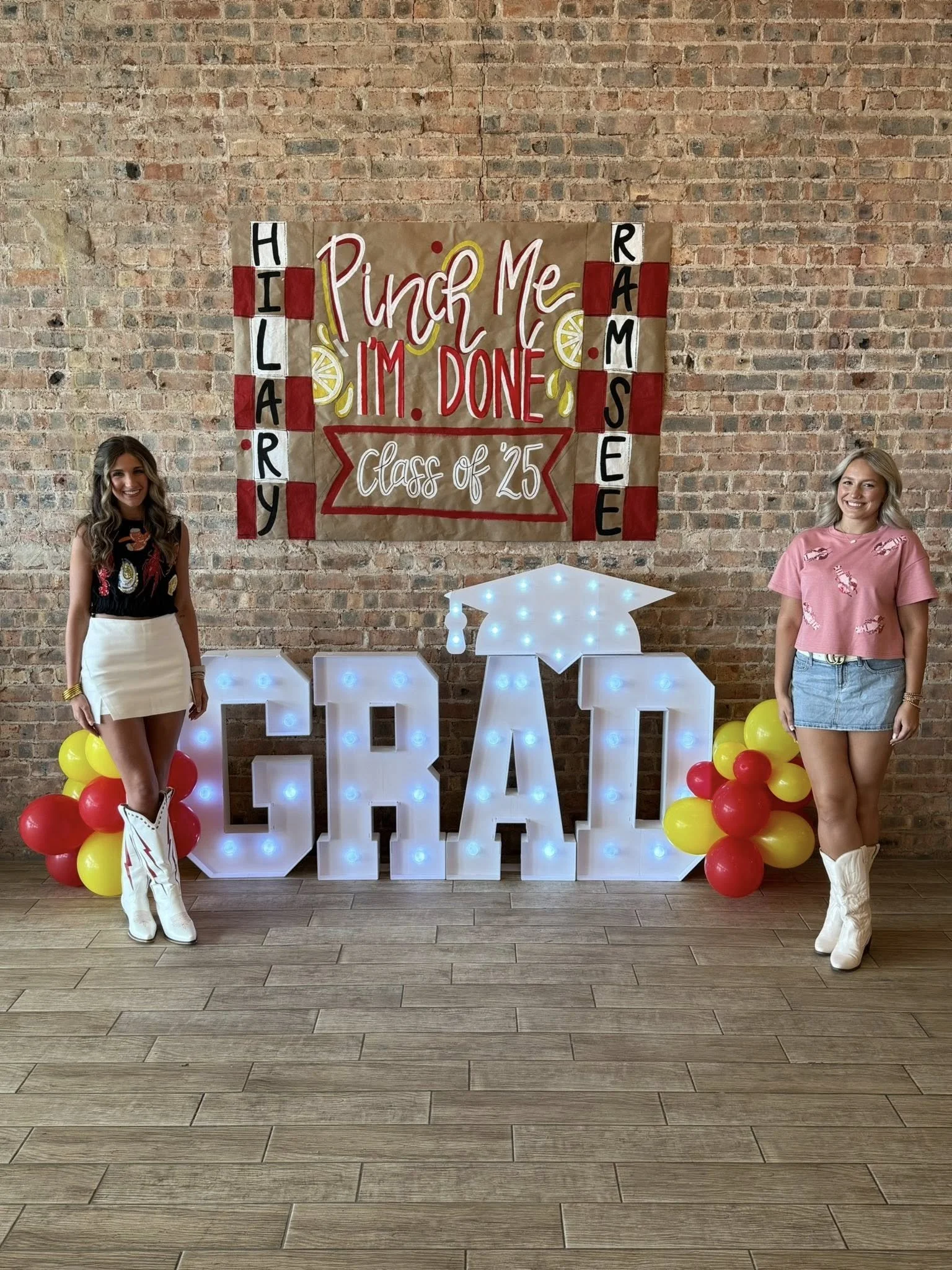 Graduation celebration with two young women in front of a brick wall. Decor includes large illuminated letters spelling 'GRAD', balloons in red and yellow, and a large paper sign with a checkered pattern, lemon slices, and the text 'Pinch Me, I'm Don