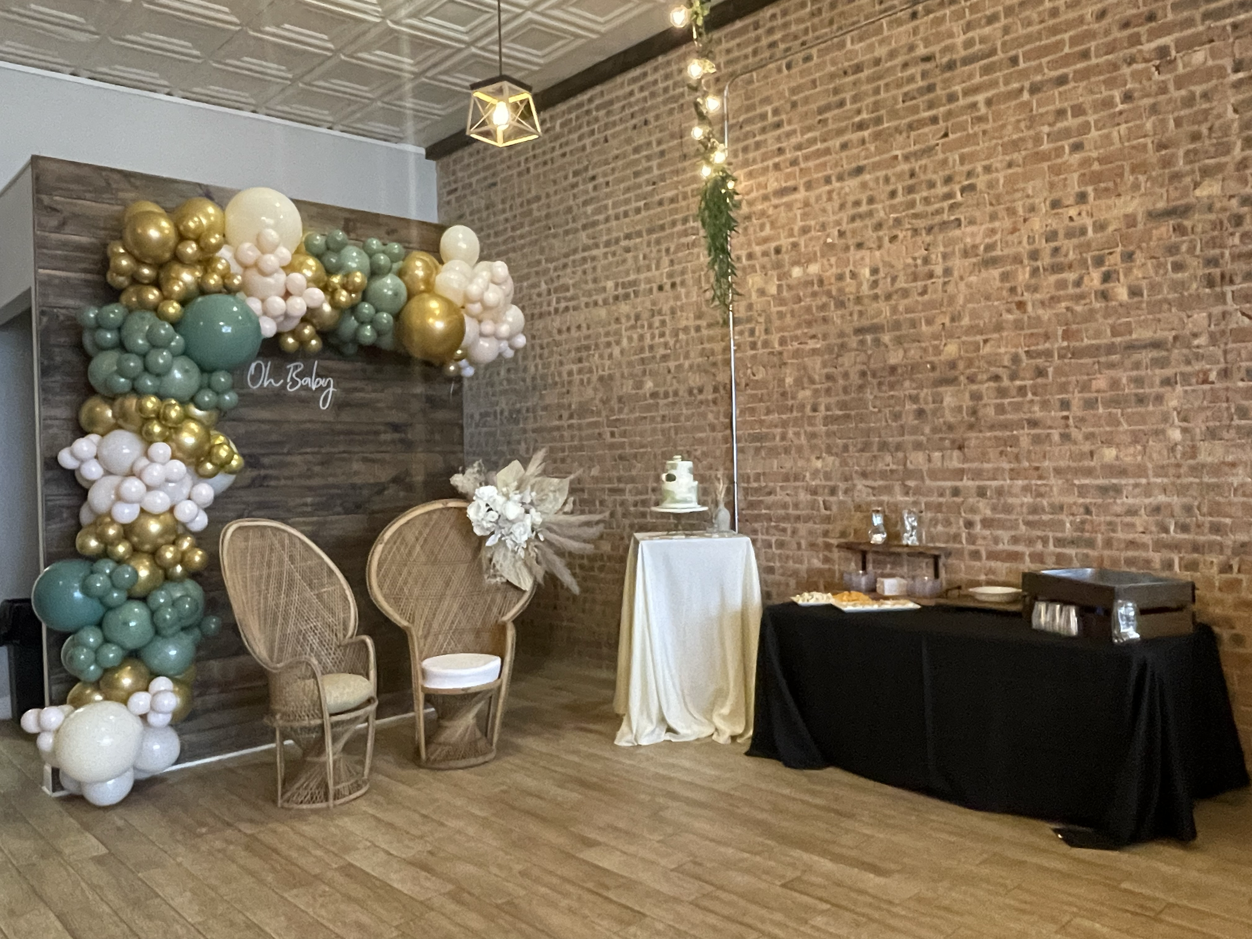 Decorative event setup with a balloon arch, two woven chairs, a table with a floral arrangement, a cake on a stand, and a buffet table, against a brick wall in a rustic indoor space.