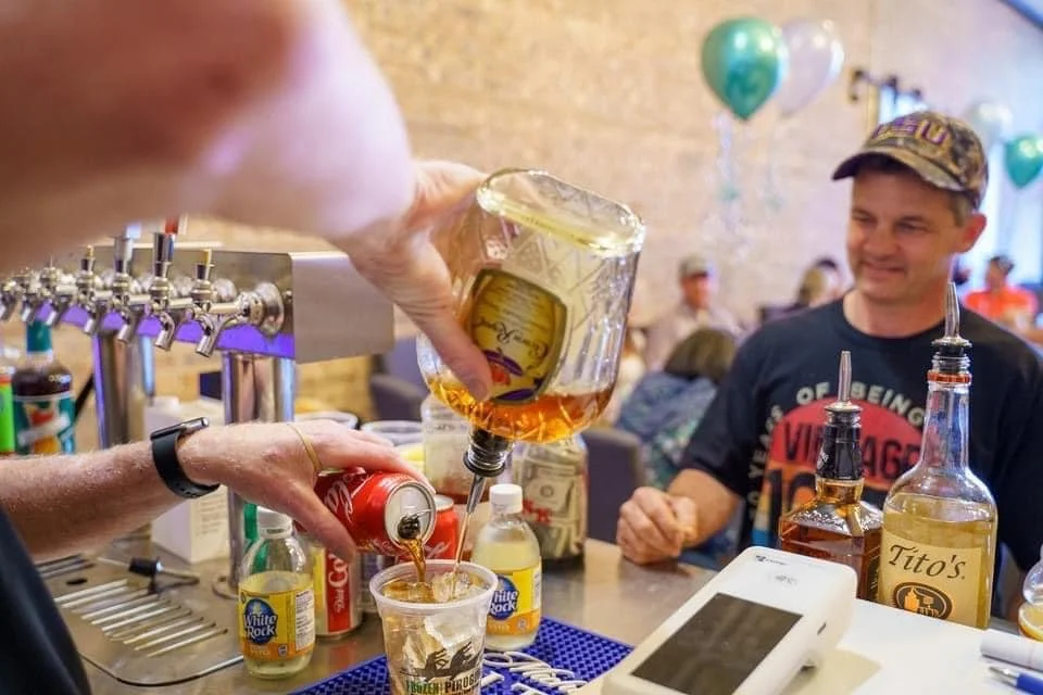 A person pouring beer from a large glass beer pitcher into a plastic cup at a bar, with a smiling man in a black t-shirt and camouflage baseball cap in the background.
