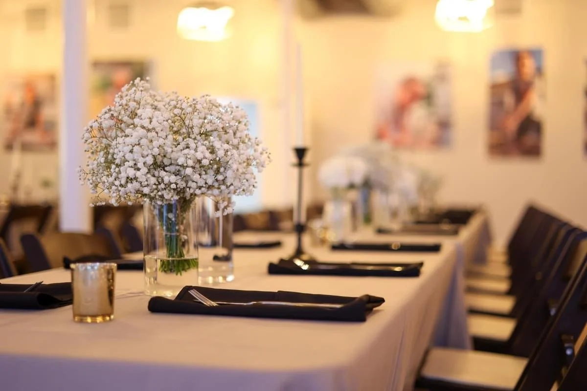 Elegant banquet table set with black napkins and silverware, decorated with white floral centerpieces in glass vases, and lit by candleholders in a softly lit, decorated room.