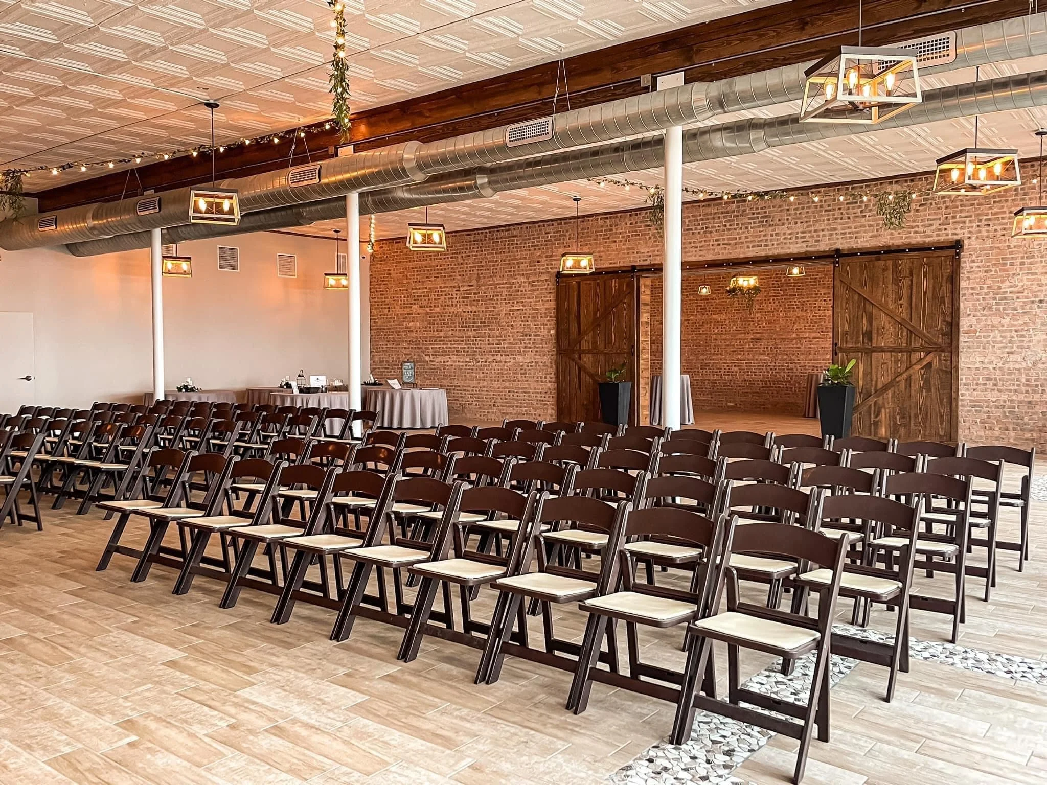 An empty event space with rows of black chairs with white cushions arranged for a gathering. There are large wooden barn doors at the back and decorative ceiling tiles. The room features exposed brick walls, potted plants, and modern pendant lighting
