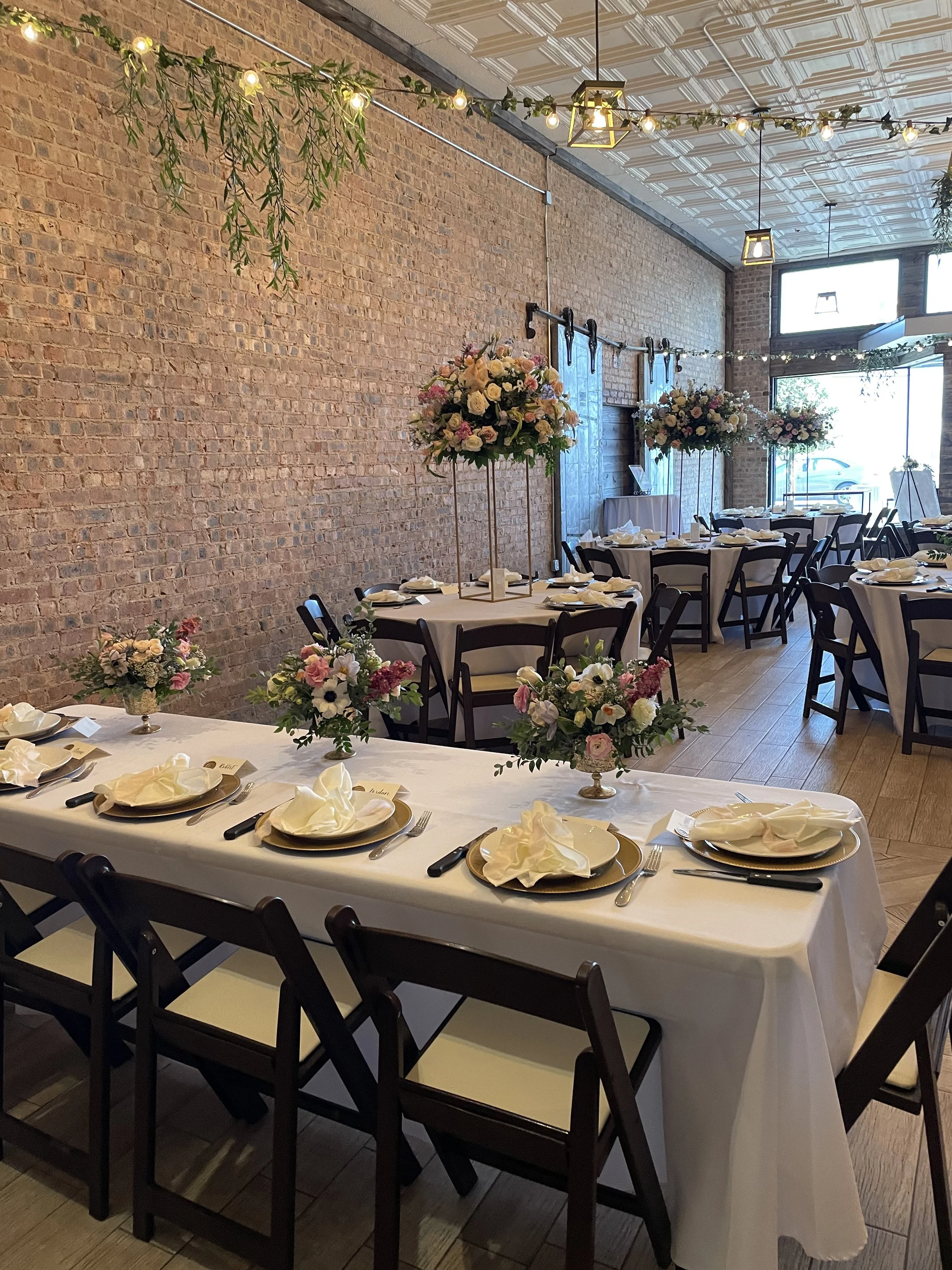 Elegant indoor wedding reception setup with round and rectangular tables decorated with white tablecloths, floral centerpieces featuring pink and white flowers, and place settings with napkins, plates, and cutlery. The room has a brick wall, string l