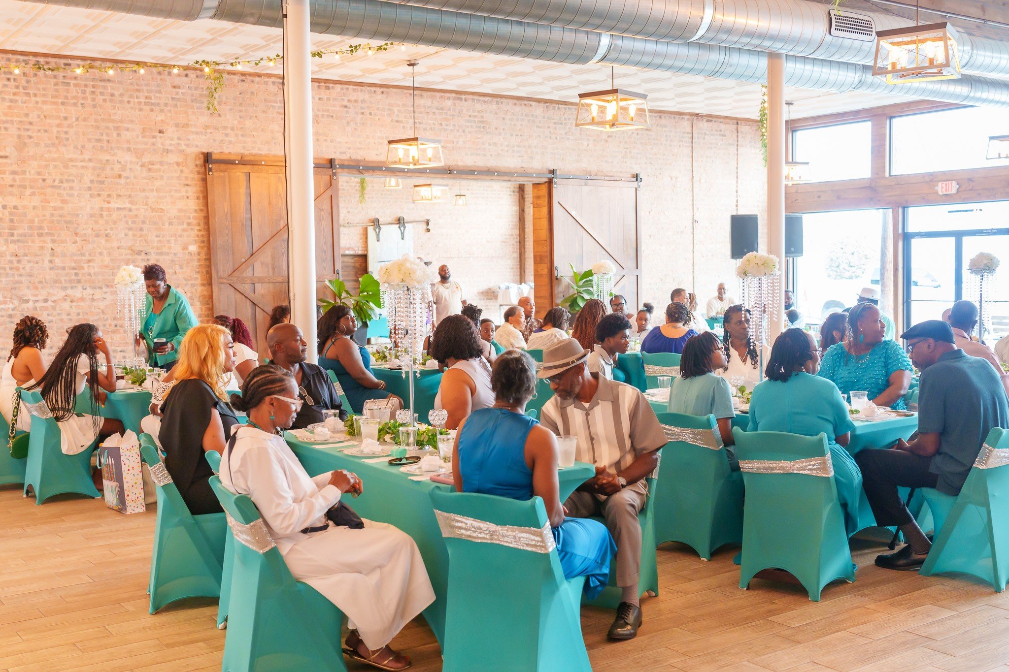 People gathered at a social event in a decorated indoor venue with teal tablecloths, flower centerpieces, and string lights, engaging in conversations.