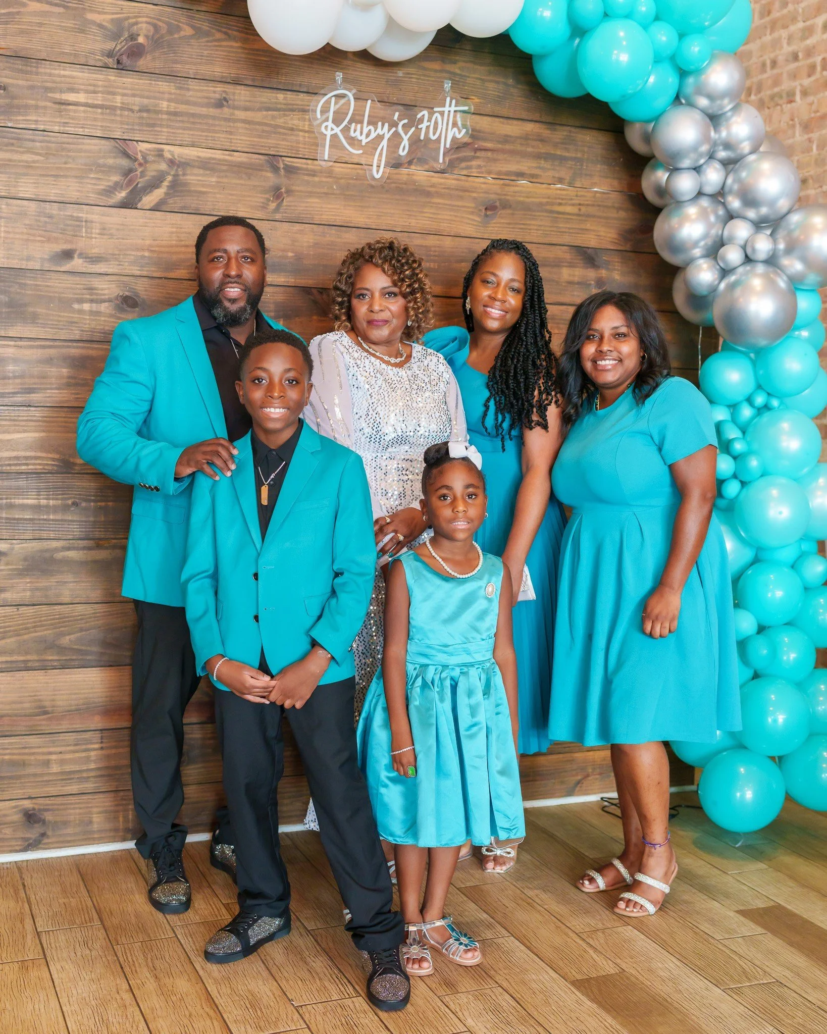 Family celebrating Ruby's 10th birthday with teal and silver decorations, including a balloon arch and a wooden backdrop. The family is dressed in teal and formal attire.