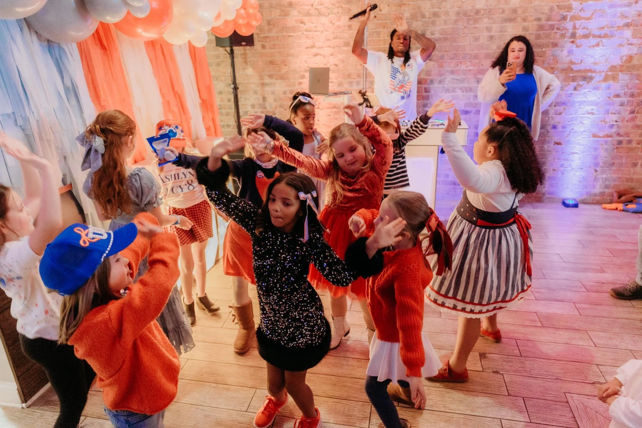 Children dancing and having fun at a birthday party with orange, white, and blue decorations, including balloons and a table in the background.