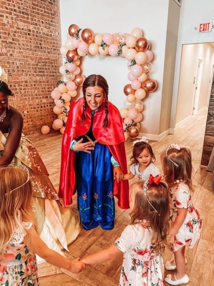A woman dressed as a princess, wearing a red cape and blue dress, is surrounded by young girls holding hands and smiling. The scene is set at a celebration with pink and gold balloons arranged in an arch in the background.