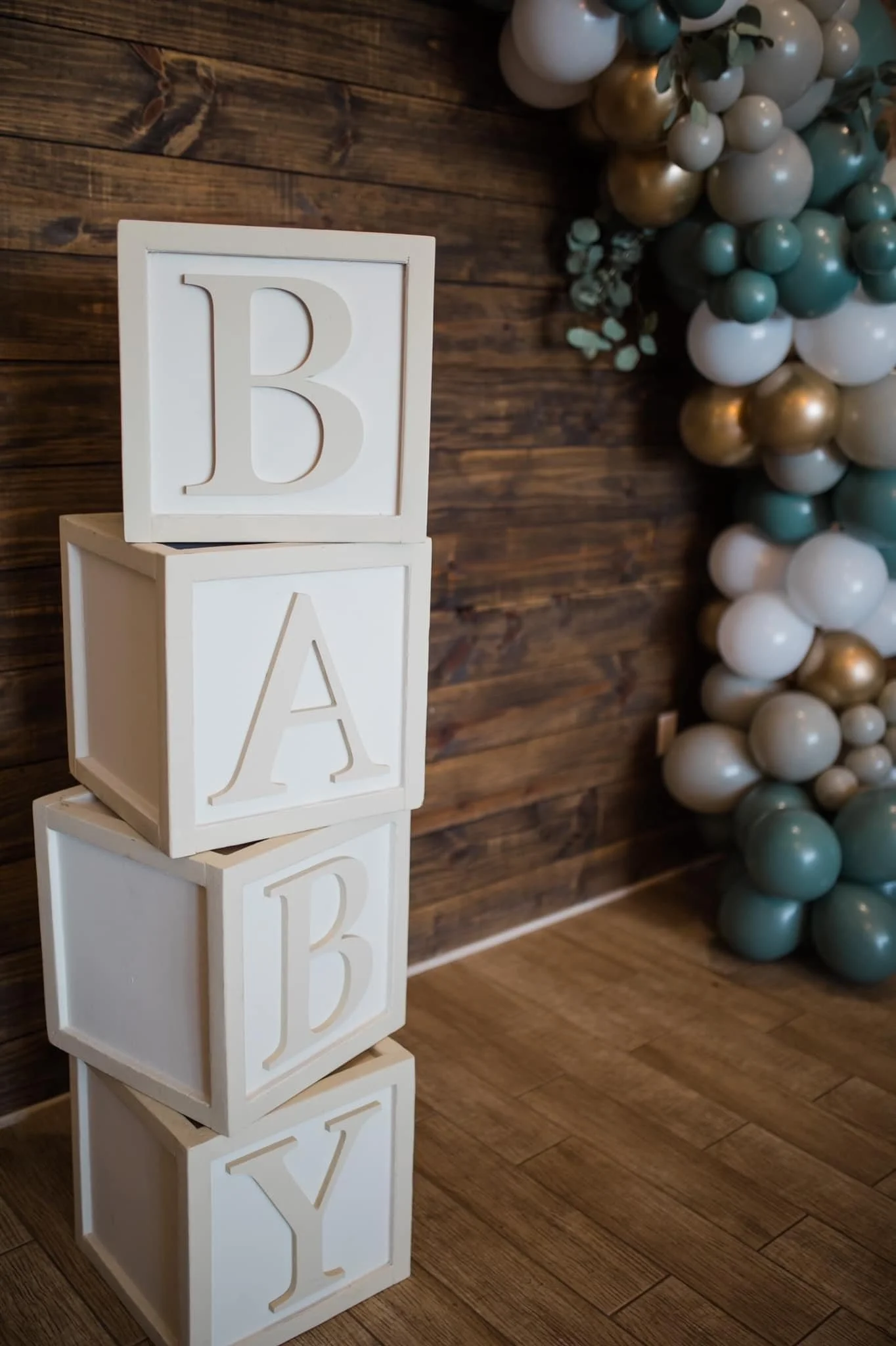 A vertical stack of four white wooden blocks with embossed letters B, A, B, Y on each side, arranged on a wooden floor against a dark wooden wall, with a balloon decoration in shades of white, gold, and blue in the background.