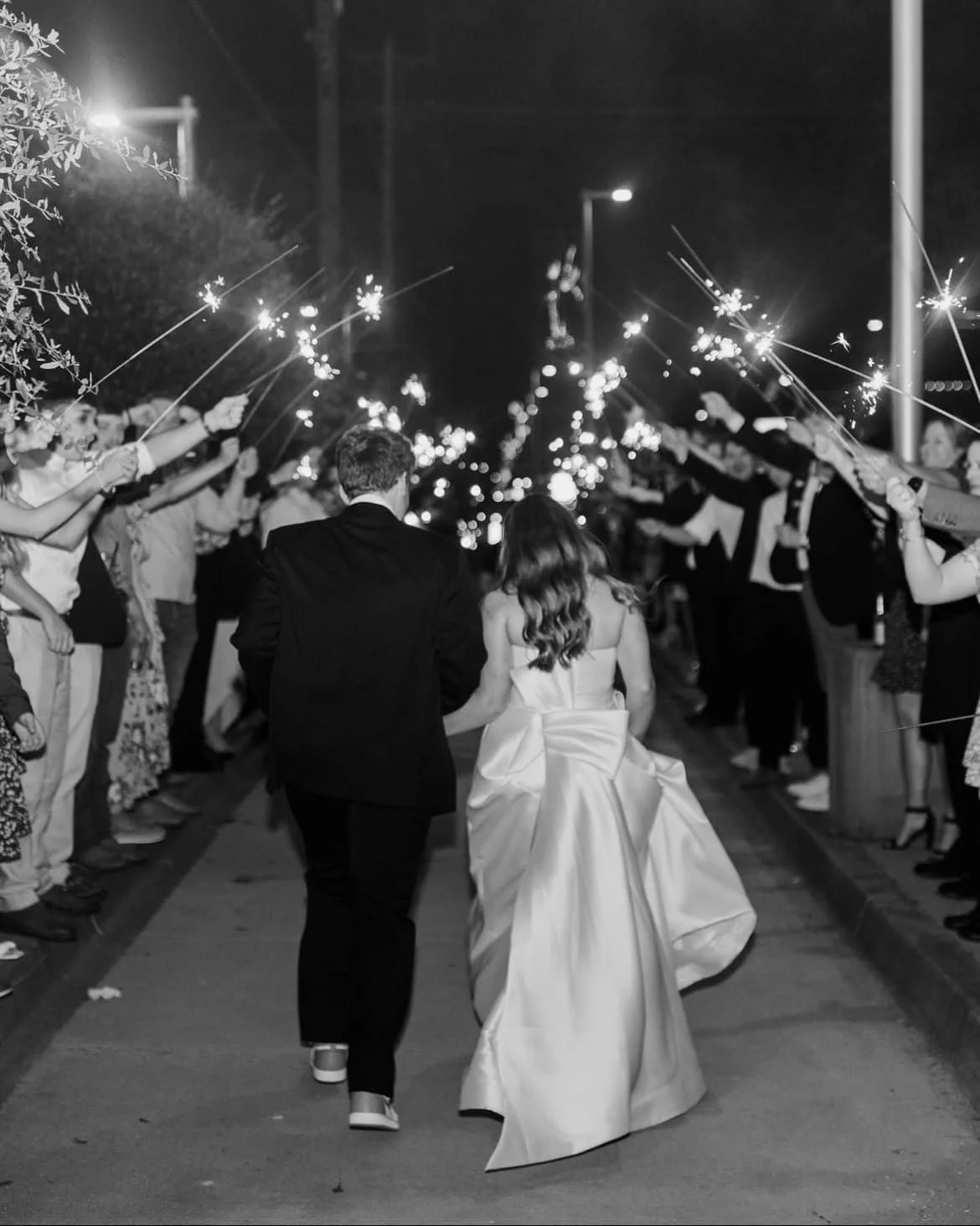 A black and white photo of a couple walking hand in hand through a crowd at night, holding sparklers, during a celebration or wedding event.