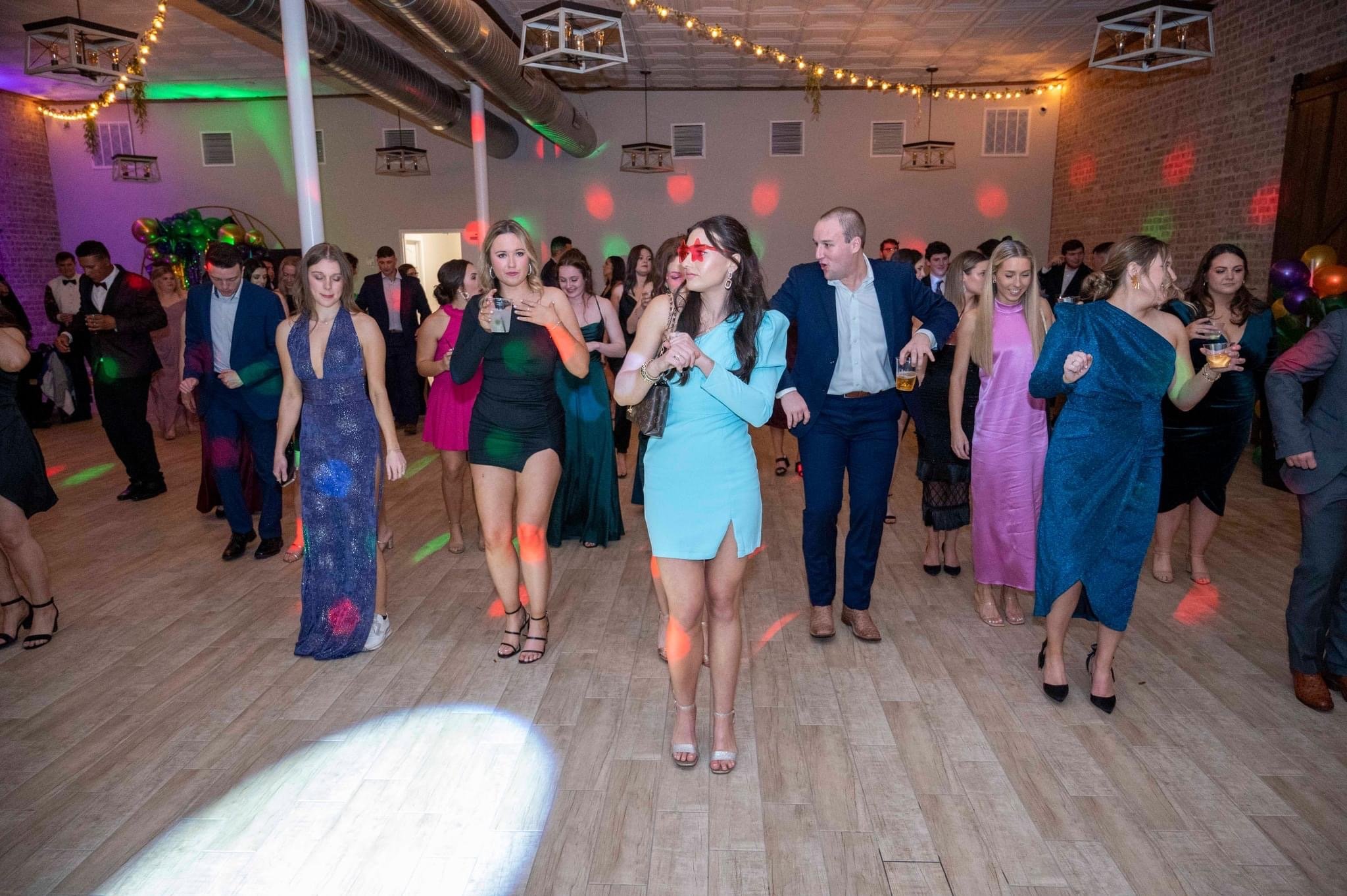 People dancing at a party or celebration in a decorated hall with wooden floors and string lights.