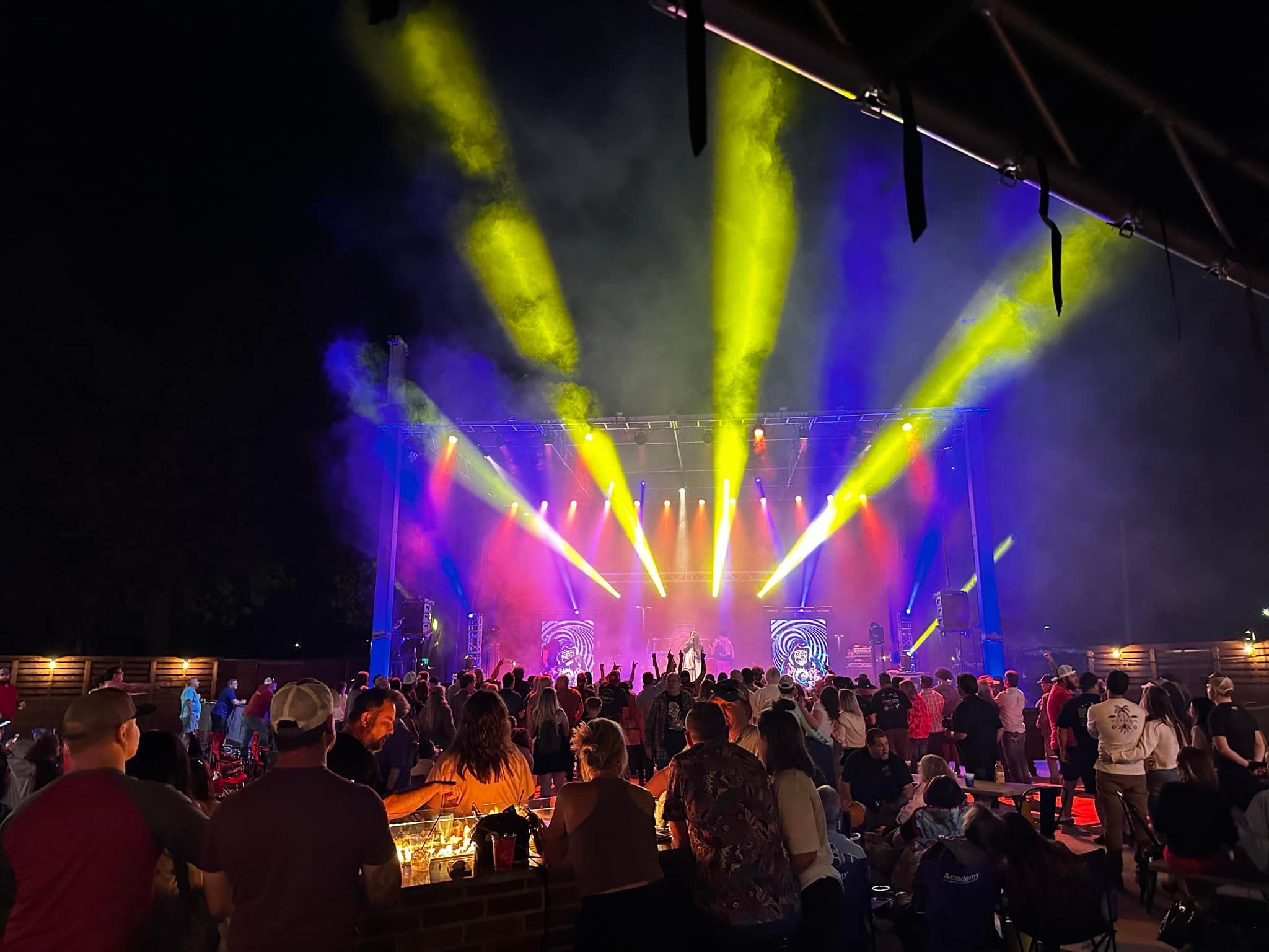 Nighttime outdoor concert with a stage illuminated by yellow, blue, and purple lights, crowd of people enjoying the performance.