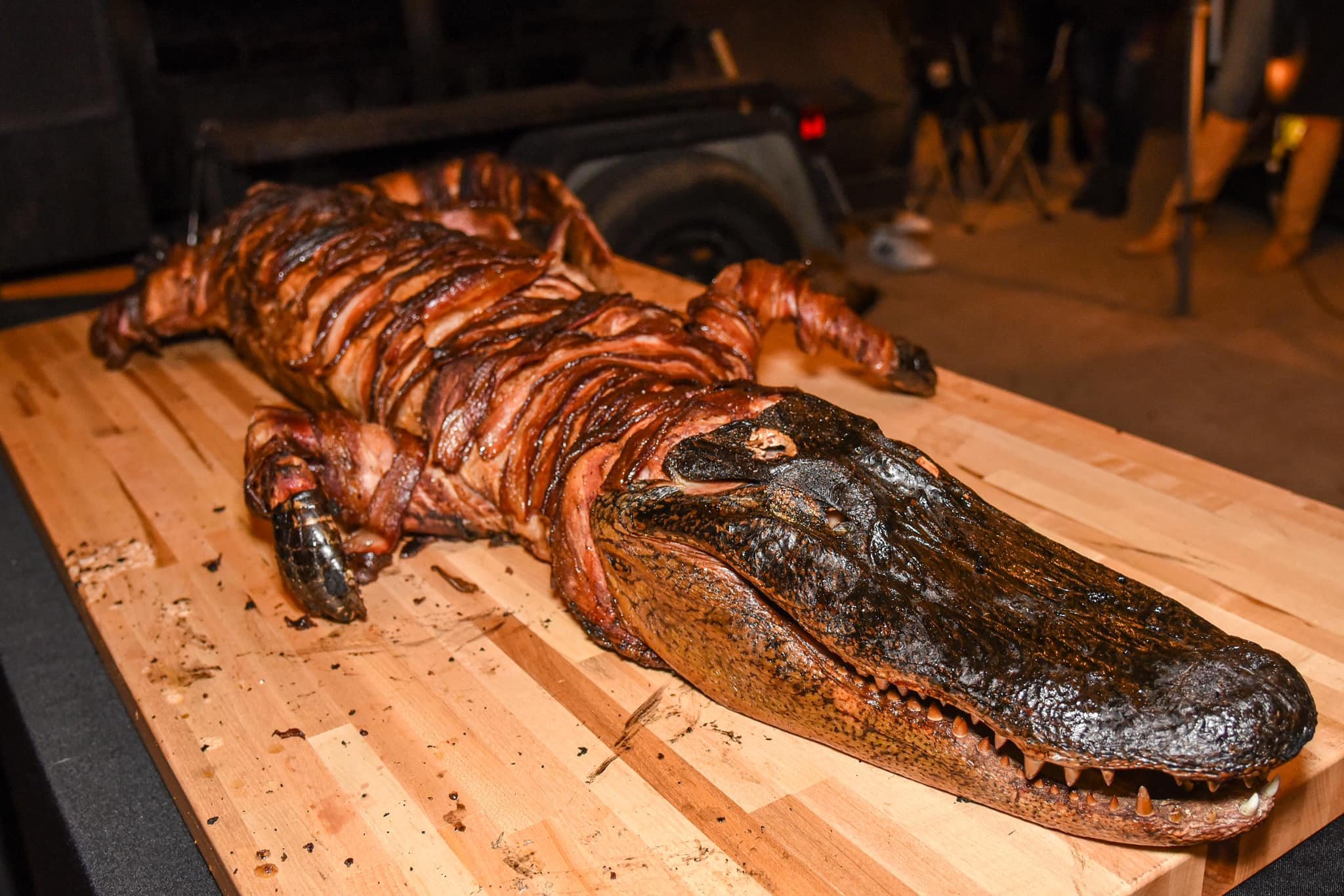 A whole roasted crocodile on a wooden table, with its head and tail intact, skin dark and shiny, and a series of sliced segments along its body.