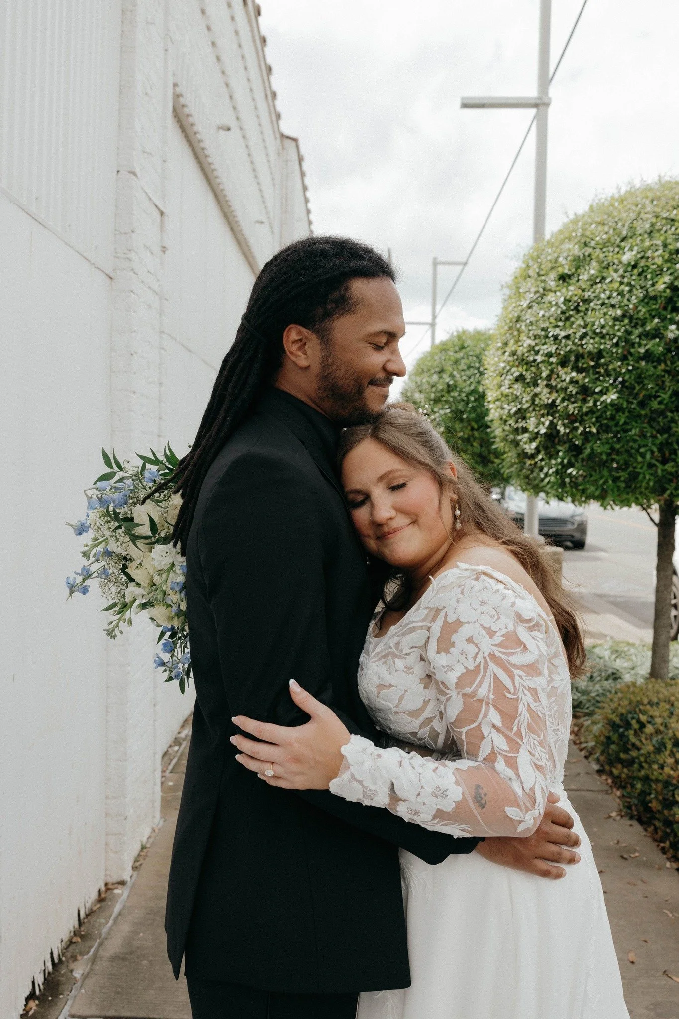 A couple dressed in wedding attire embracing each other on a sidewalk, with greenery and a street in the background.