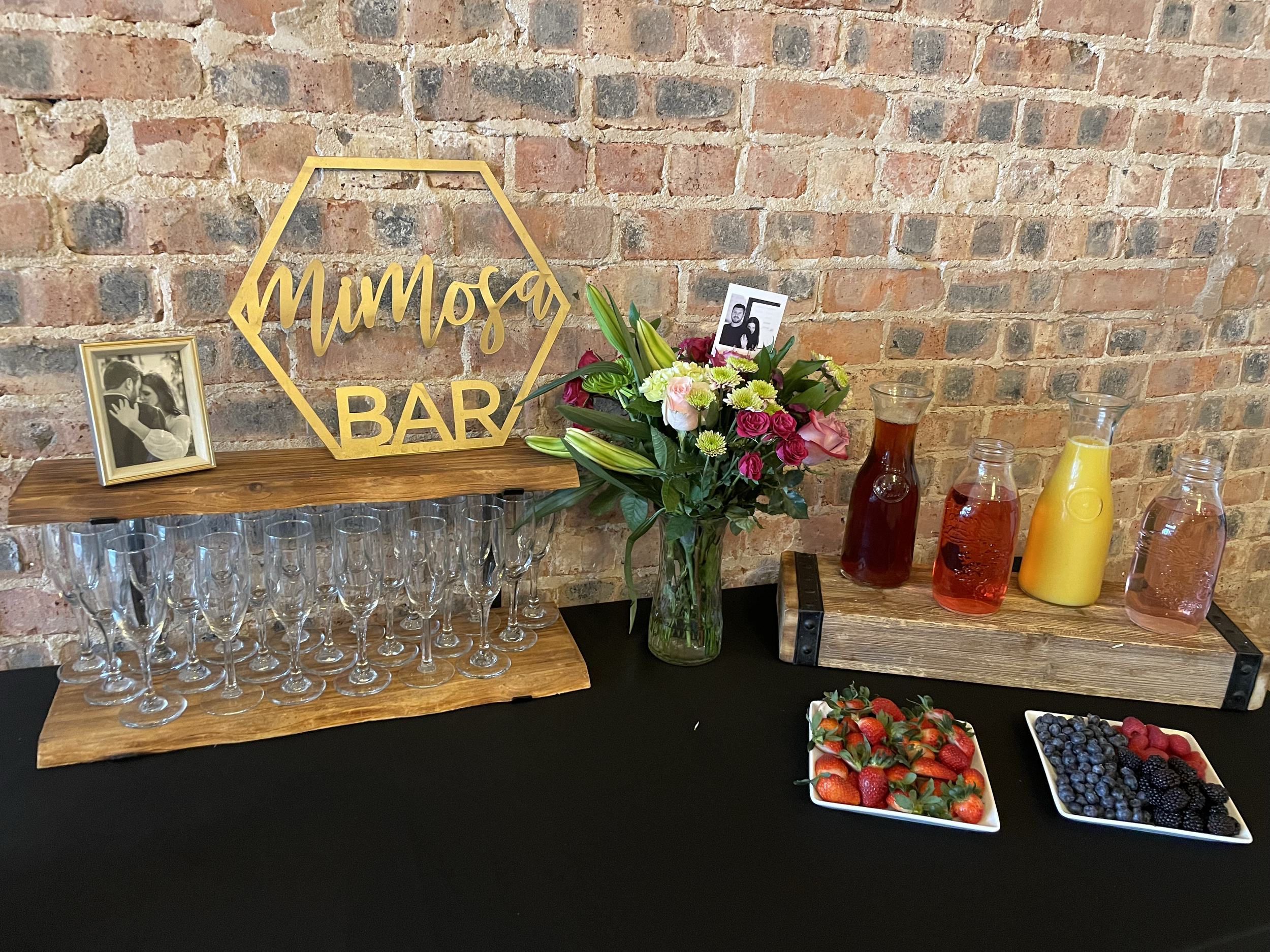 Bar setup with glasses, flowers, fruit, and beverages against a brick wall.