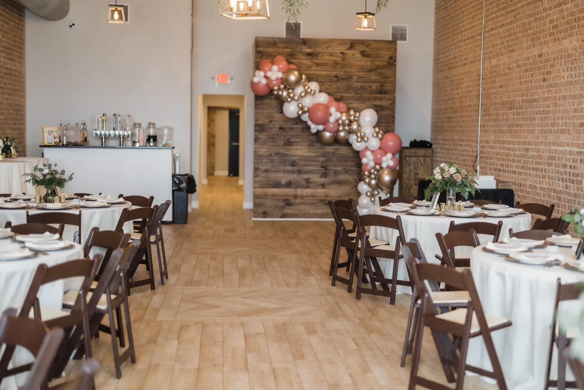 Event space decorated with round tables covered with white tablecloths and wooden chairs, floral centerpieces, and balloon arch in pink, white, and gold on wooden wall backdrop.