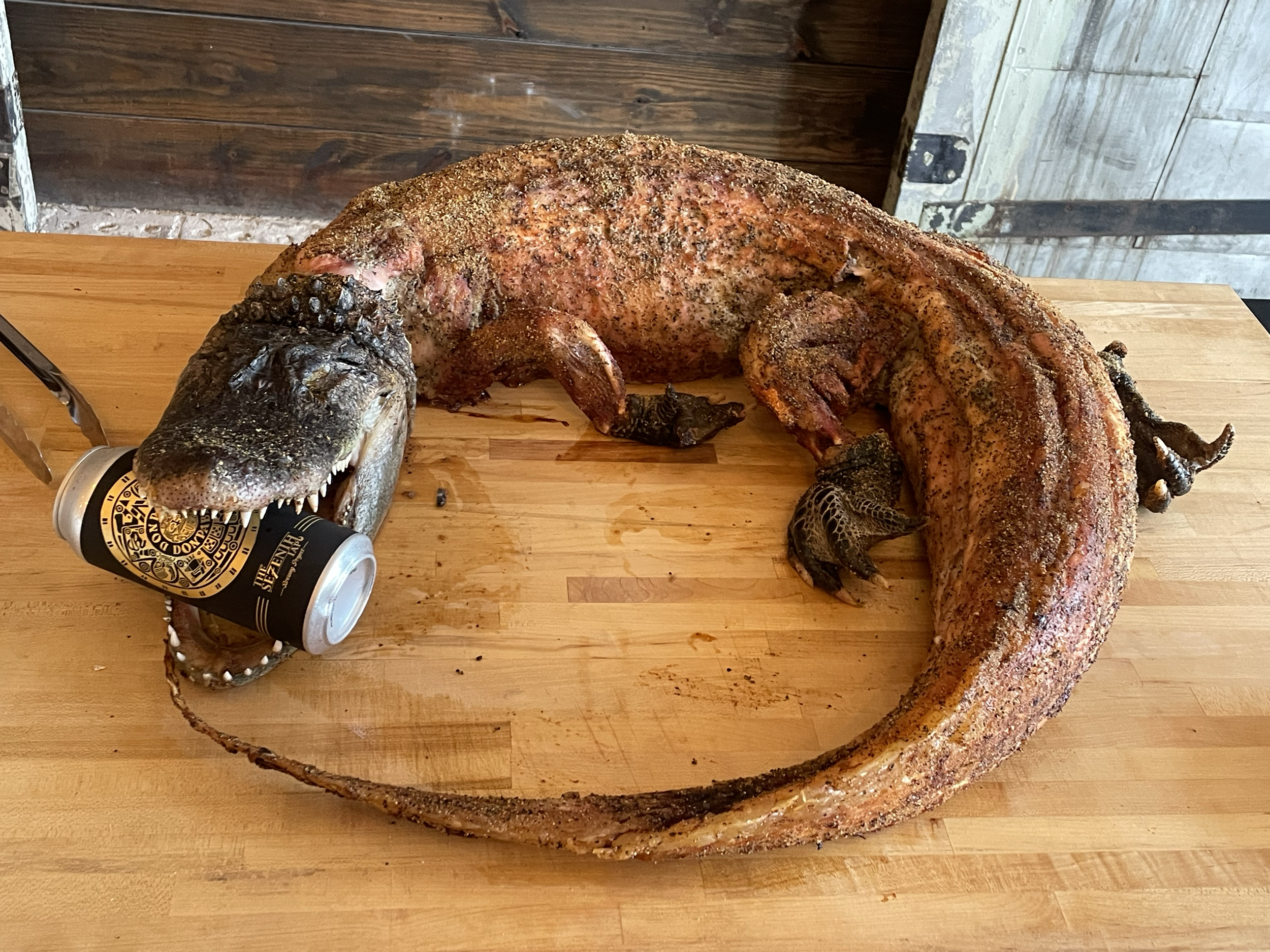 A cooked whole lizard on a wooden cutting board, holding a can of beer in its mouth.