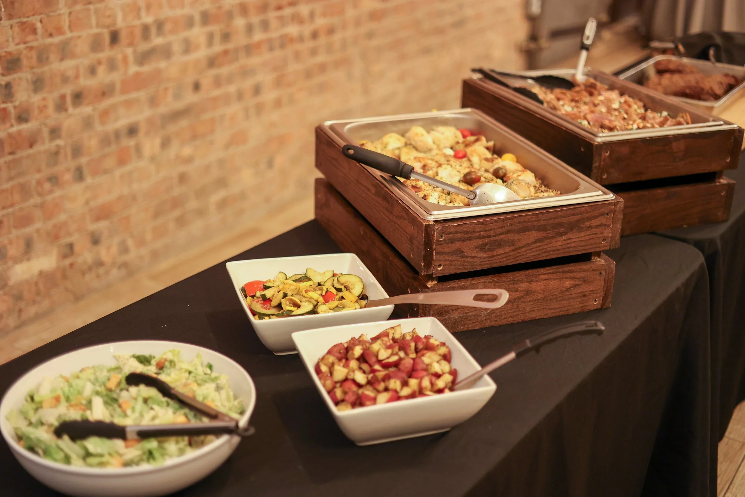 Buffet table with dishes of vegetables and cooked meats, including zucchini, red peppers, and potato salad, against a brick wall.