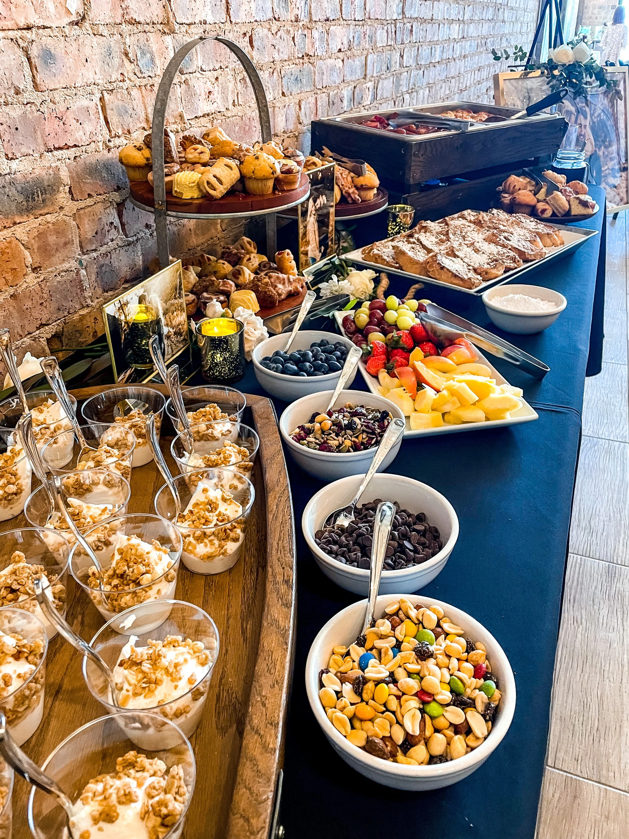 A dessert and fruit buffet table with yogurt parfaits topped with granola, bowls of blueberries, mixed berries, chocolate chips, and trail mix, along with various pastries, fruit, and a waffle station, set against an exposed brick wall.