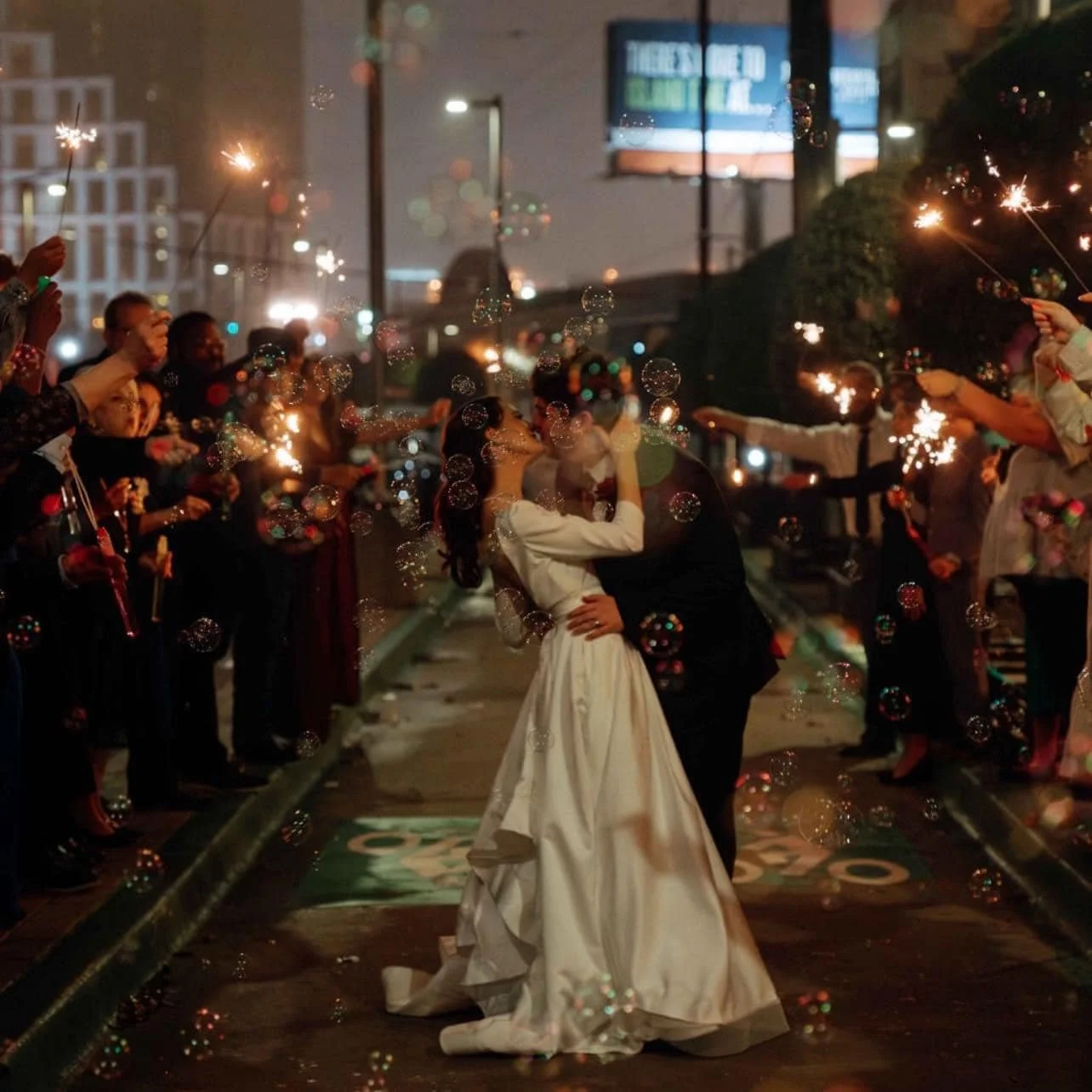 A newlywed couple sharing a kiss and dance in the streets at night, surrounded by friends and family holding sparklers, with bubbles and city lights in the background.