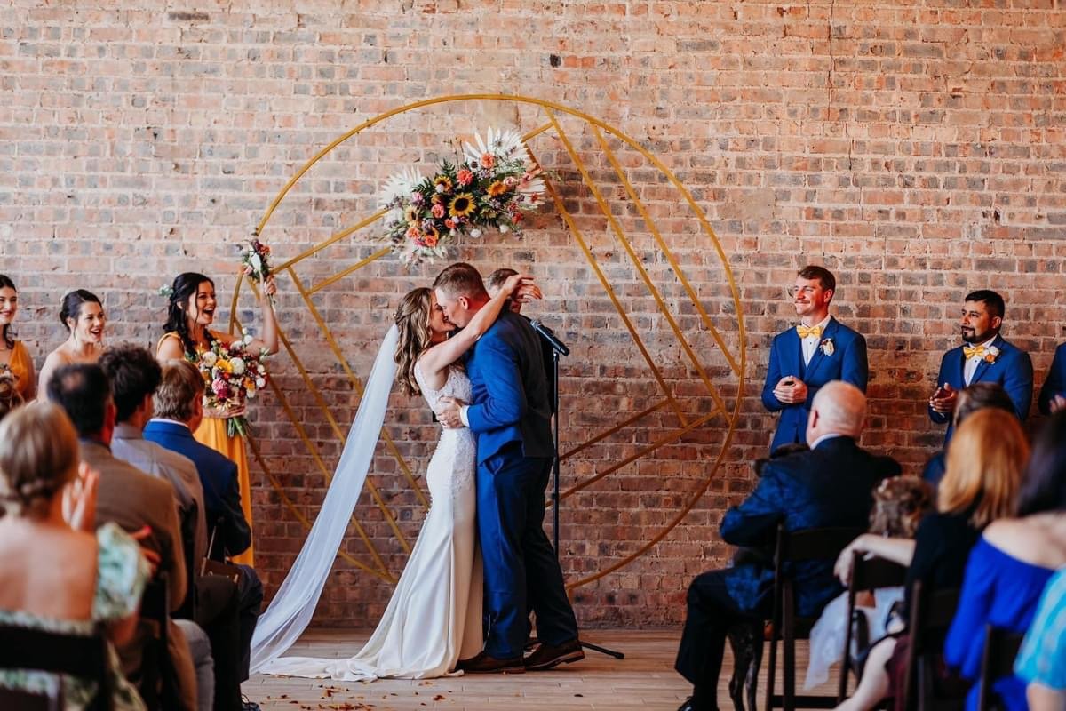 Bride and groom kissing during wedding ceremony with bridesmaids and groomsmen standing nearby, guests seated watching, brick wall backdrop with floral arrangement.