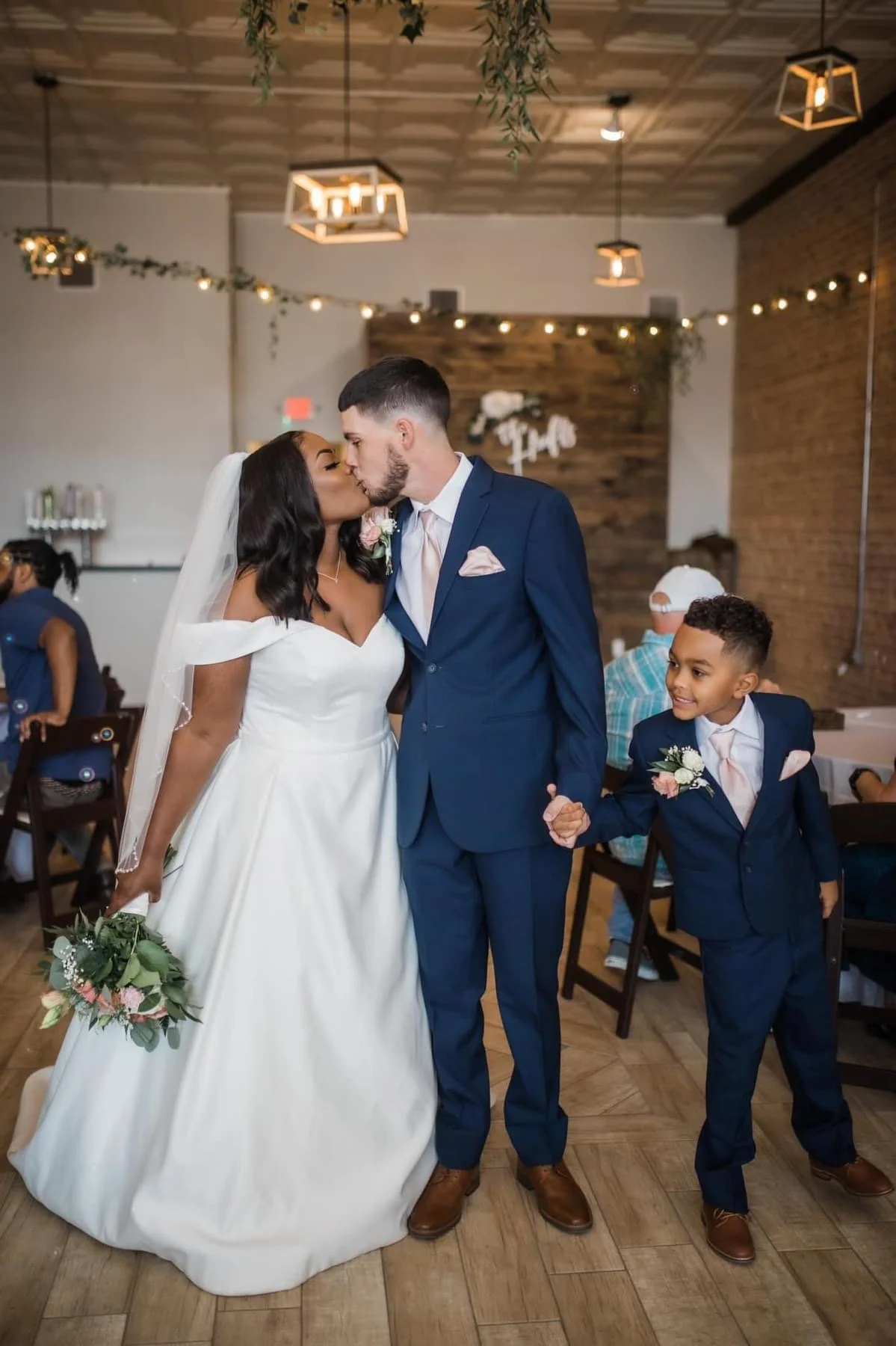 A wedding couple, a woman in a white gown and a man in a blue suit, kiss while holding hands, with a young boy in a matching blue suit holding their hands and smiling.