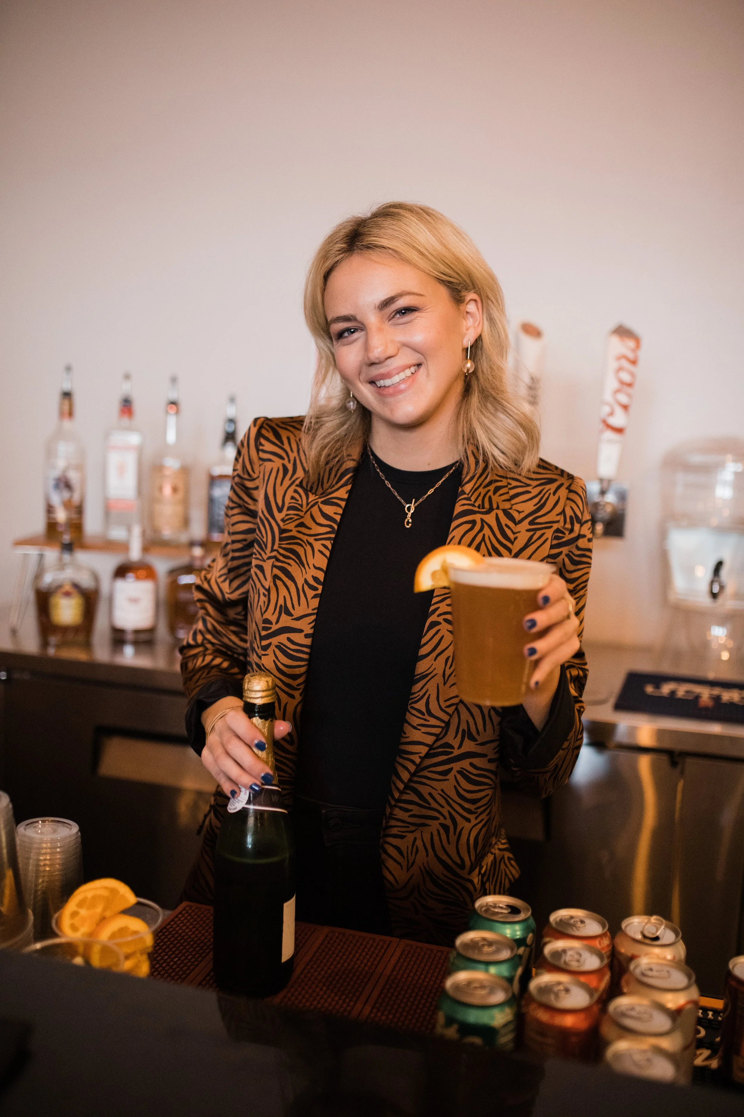 A woman with blonde hair smiling, wearing a patterned blazer and black top, holding a drink with lemon wedge, standing at a bar with various bottles and cans in the background.