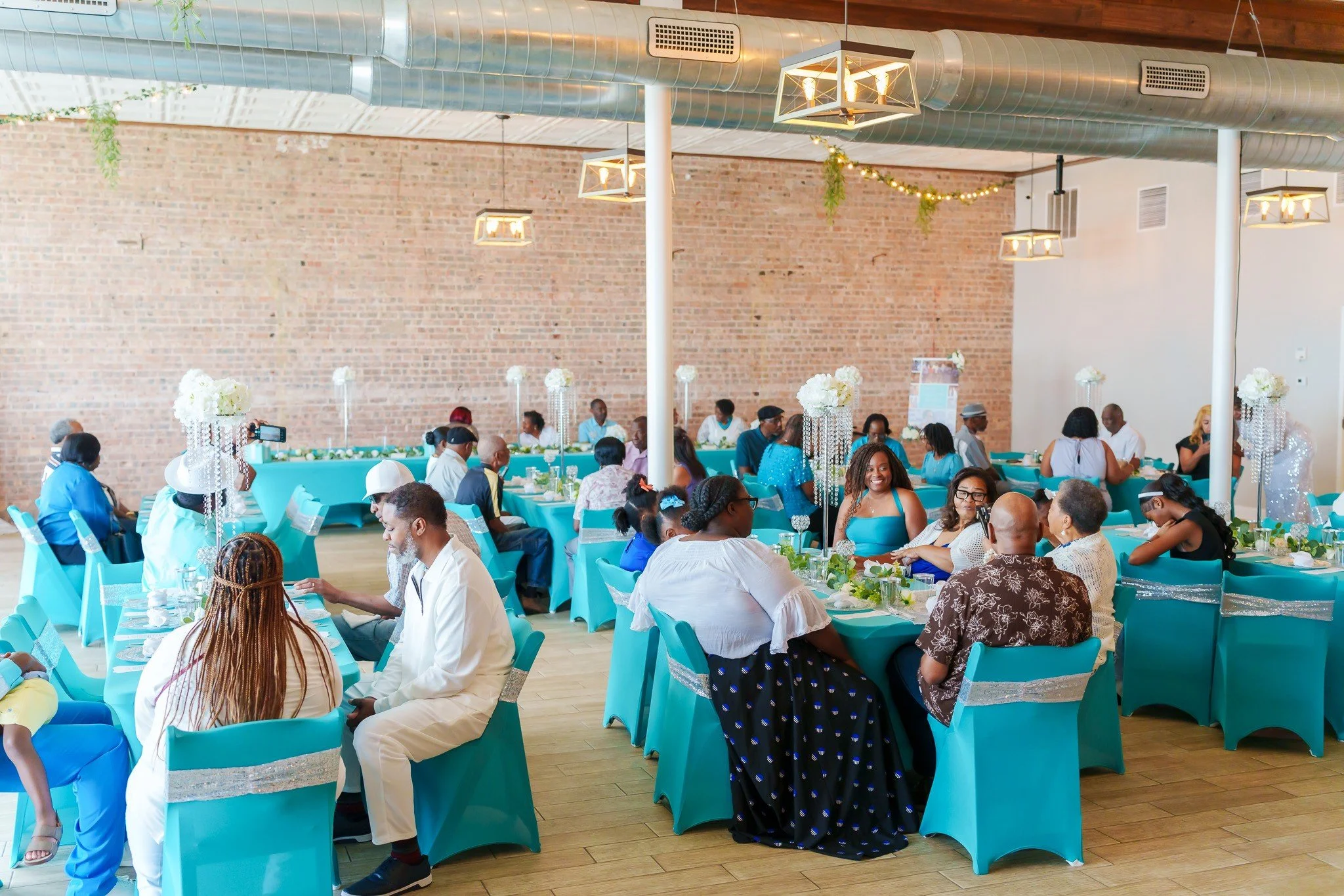 Guests at a celebration in a decorated venue with teal tablecloths and floral centerpieces, engaging in conversations and dining.