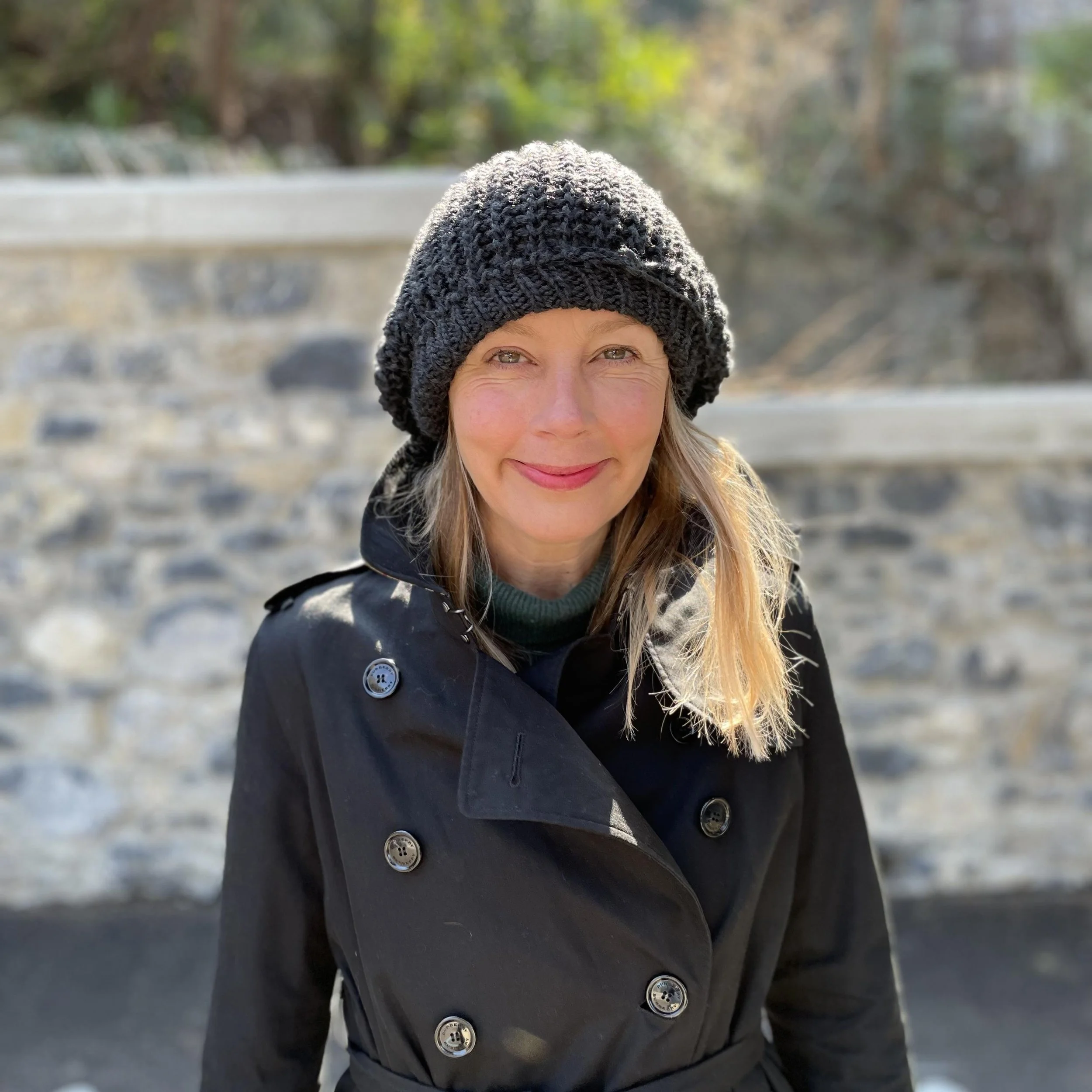A woman outdoors wearing a black coat and a black knit hat, smiling at the camera, with a stone wall and trees in the background.