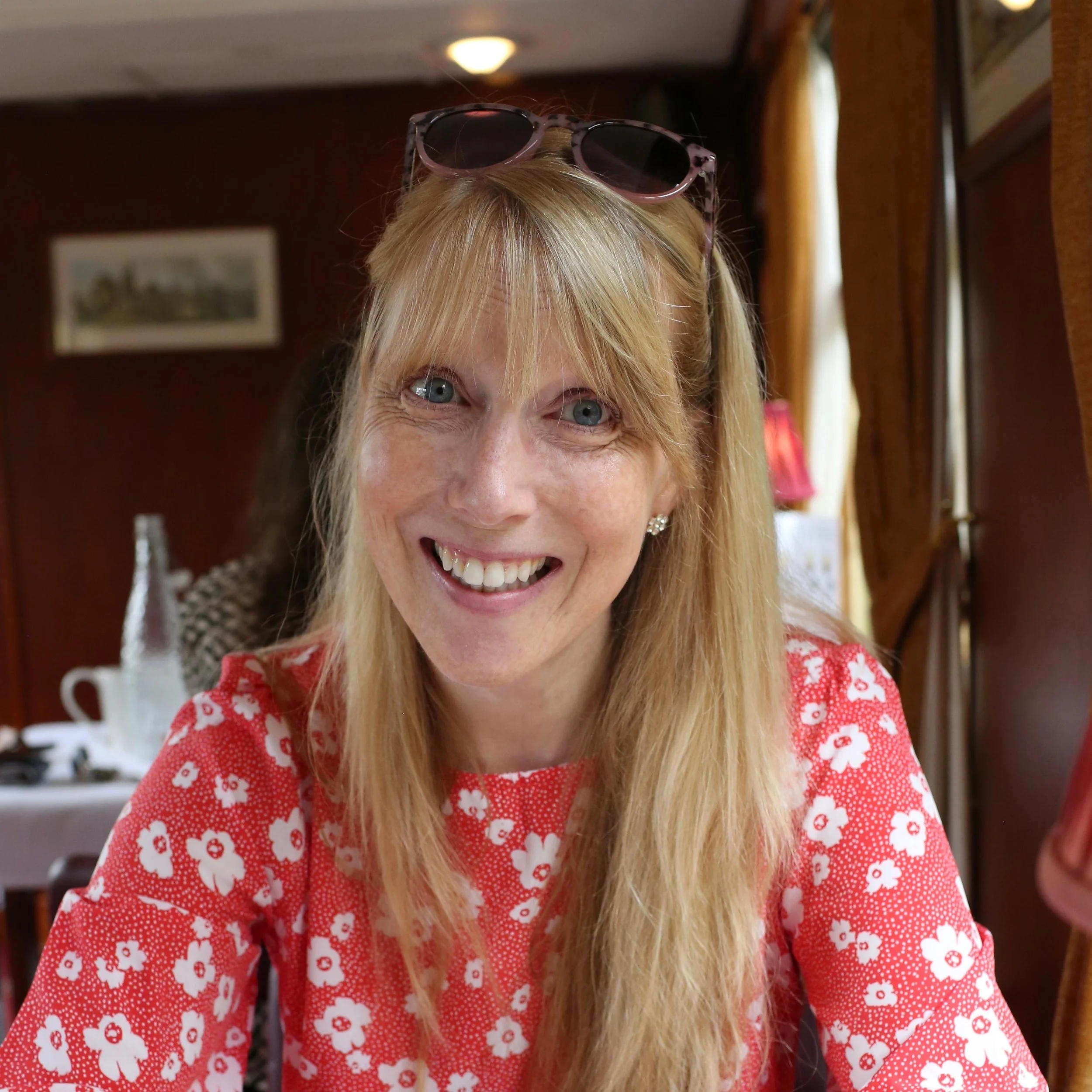 A woman with blonde hair, wearing sunglasses on her head, smiling at the camera. She is dressed in a red shirt with white floral patterns and is sitting in a cozy, warmly lit room.