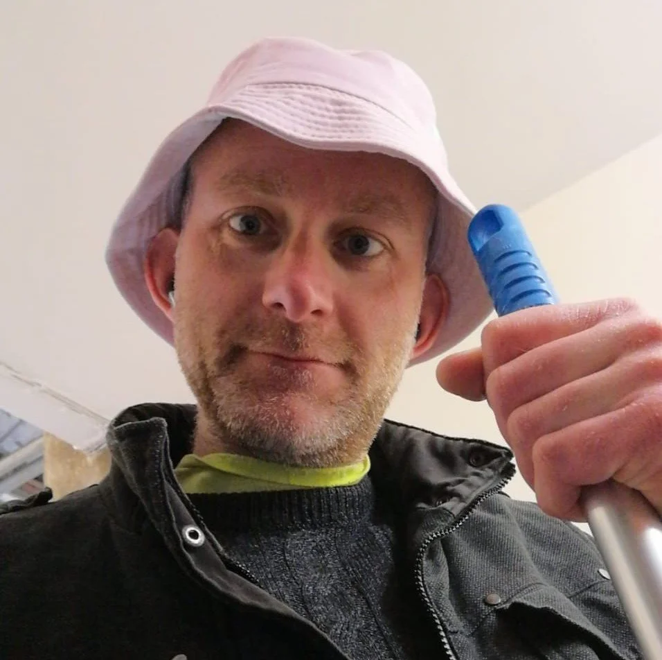 A man wearing a pink bucket hat, black jacket, and a black shirt with a yellow collar, holding a blue stick or tool