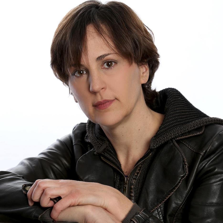 A woman with short brown hair wearing a black leather jacket, looking at the camera with a neutral expression against a white background.