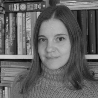A woman with long hair wearing a sweater, standing in front of a bookshelf filled with books.